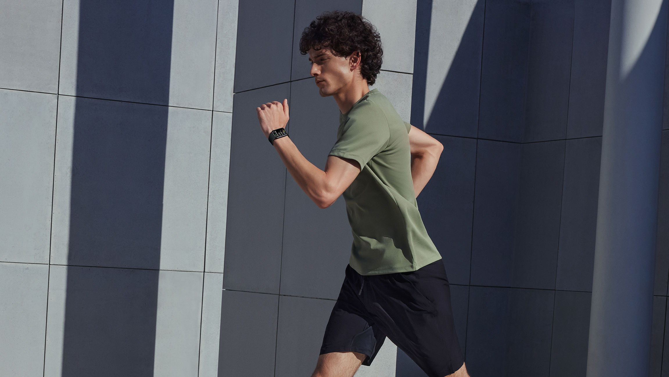 Hombre corriendo, con camisa verde y pantalones cortos negros, con un reloj inteligente.