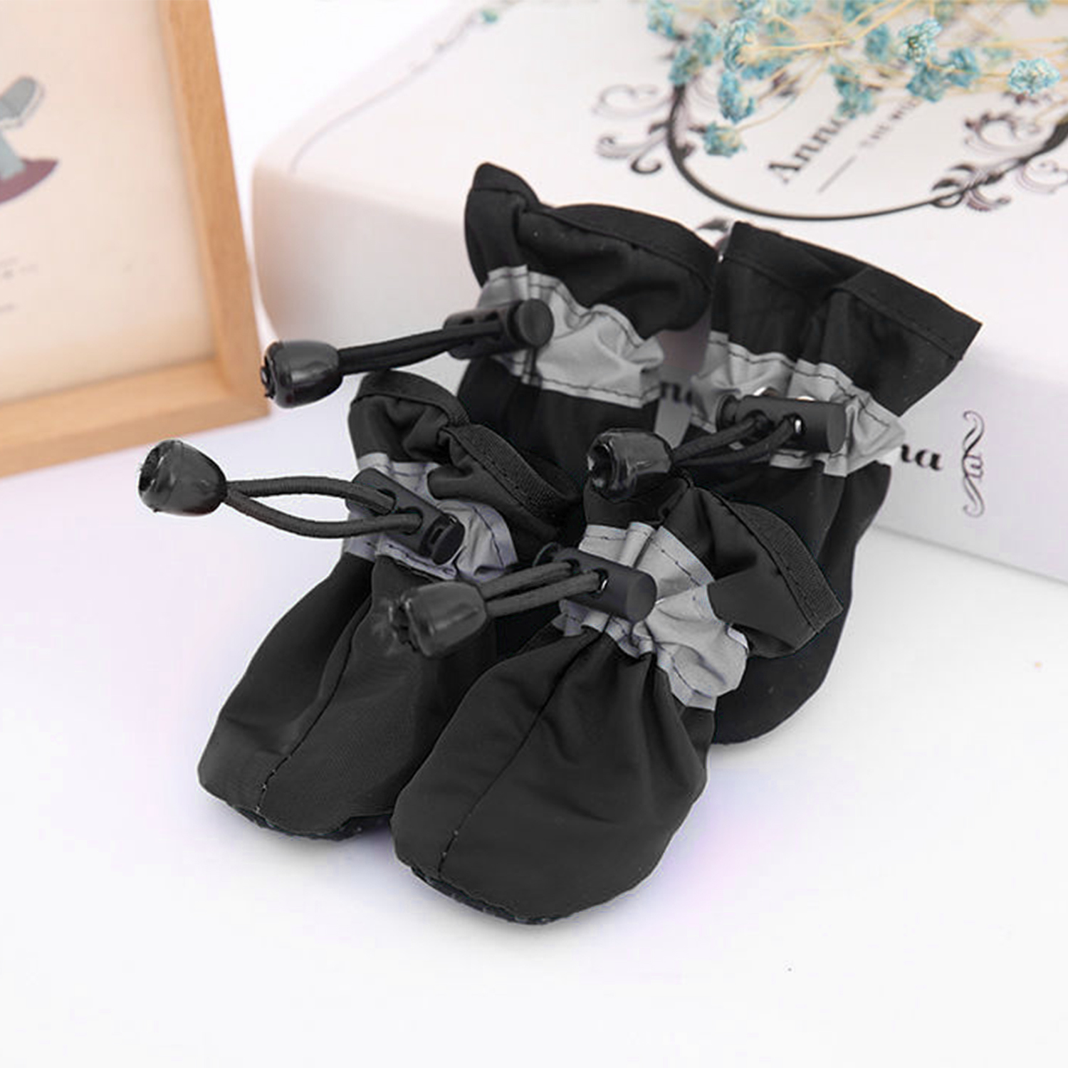 Ein Paar schwarze Hundeschuhe mit reflektierenden grauen Bändern und Kordelzügen liegen auf weißem Grund.