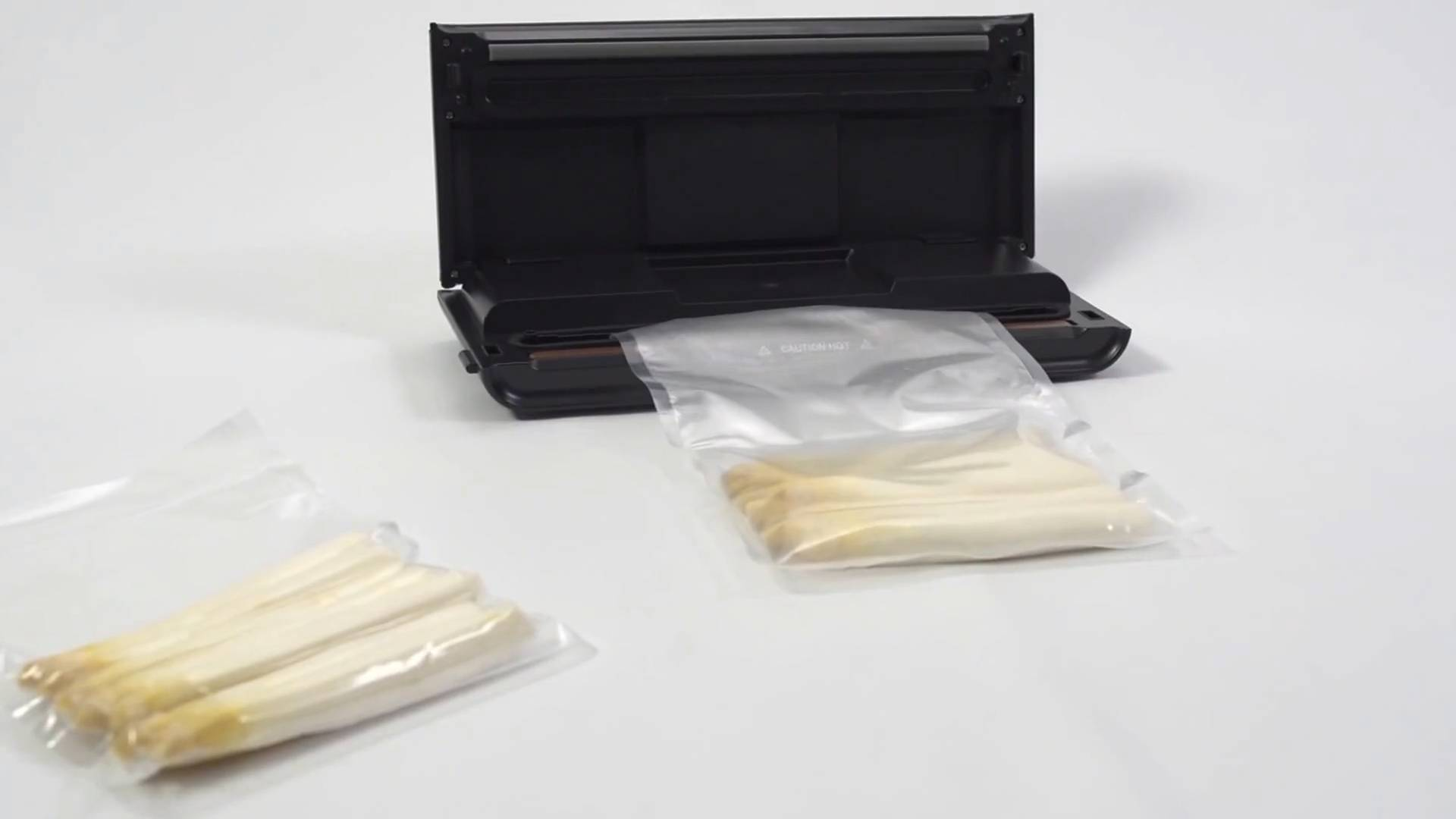 Szparagi w plastikowych torbach, obok czarnej zgrzewarki próżniowej. Białe tło.