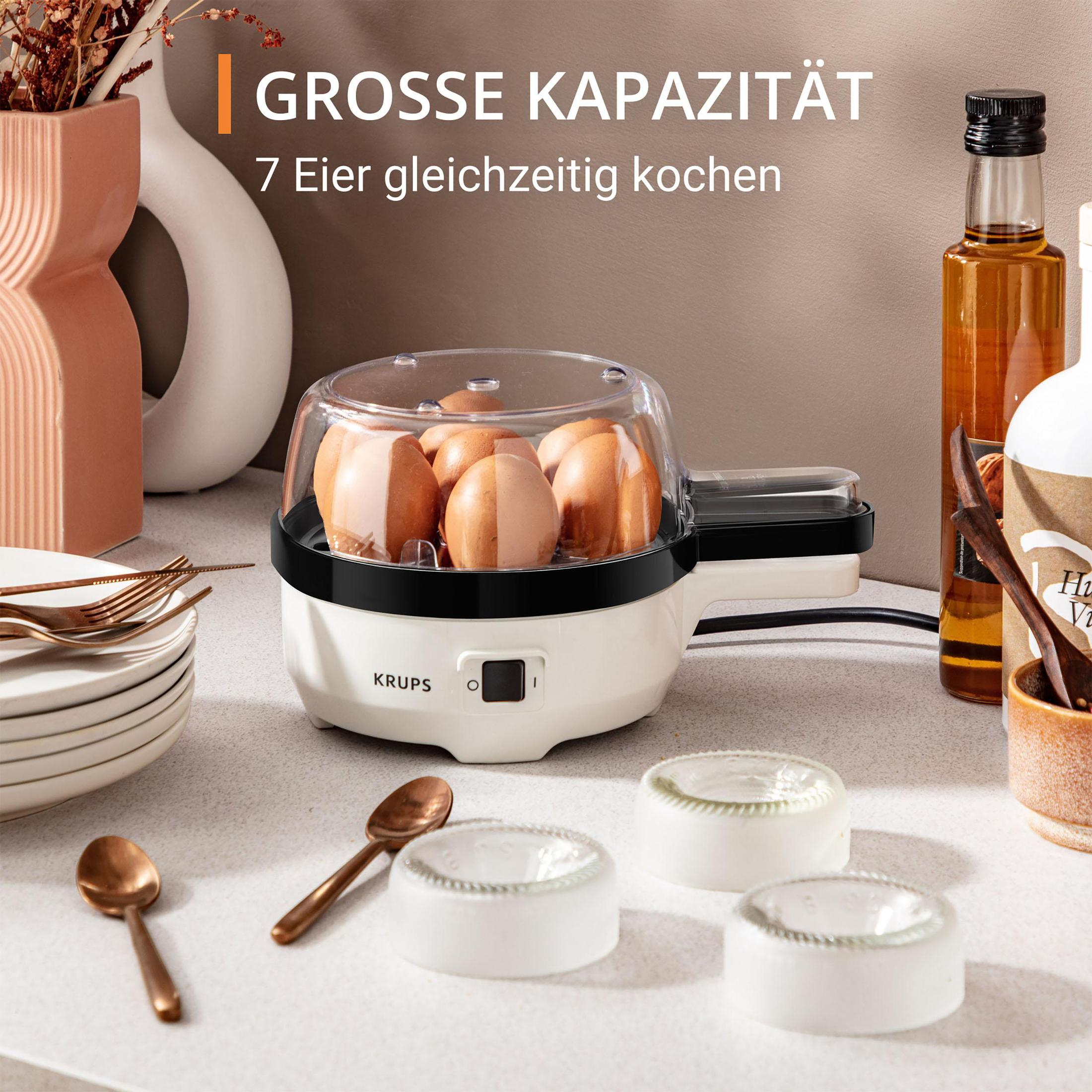 Gotowacz do jajek z 7 jajkami w środku. Gotowacz jest biały z przezroczystą pokrywką. Inne przedmioty są na blacie.