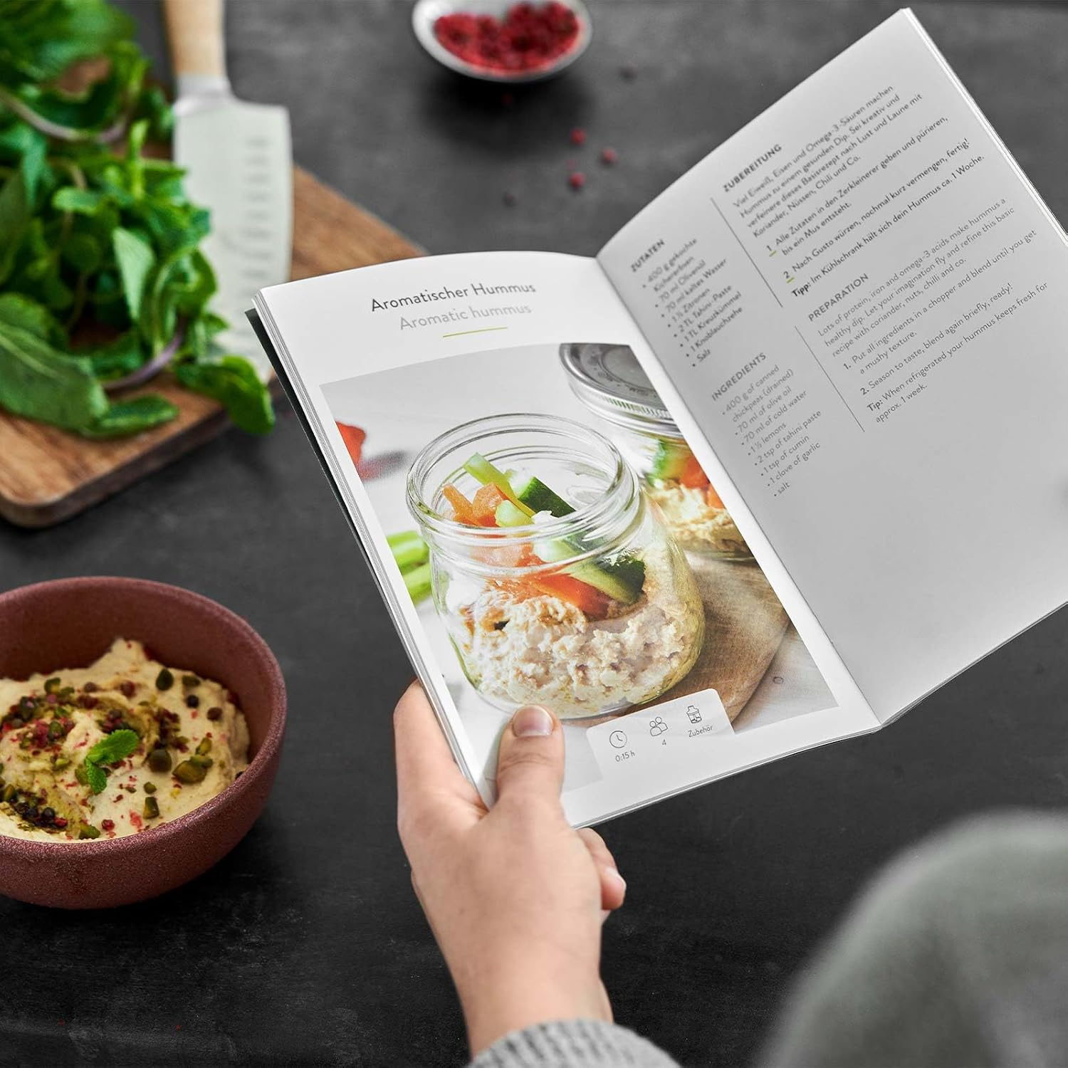 Offenes Kochbuch mit Hummus-Rezept, Hummus-Schüssel und Zutaten auf einem Tisch.