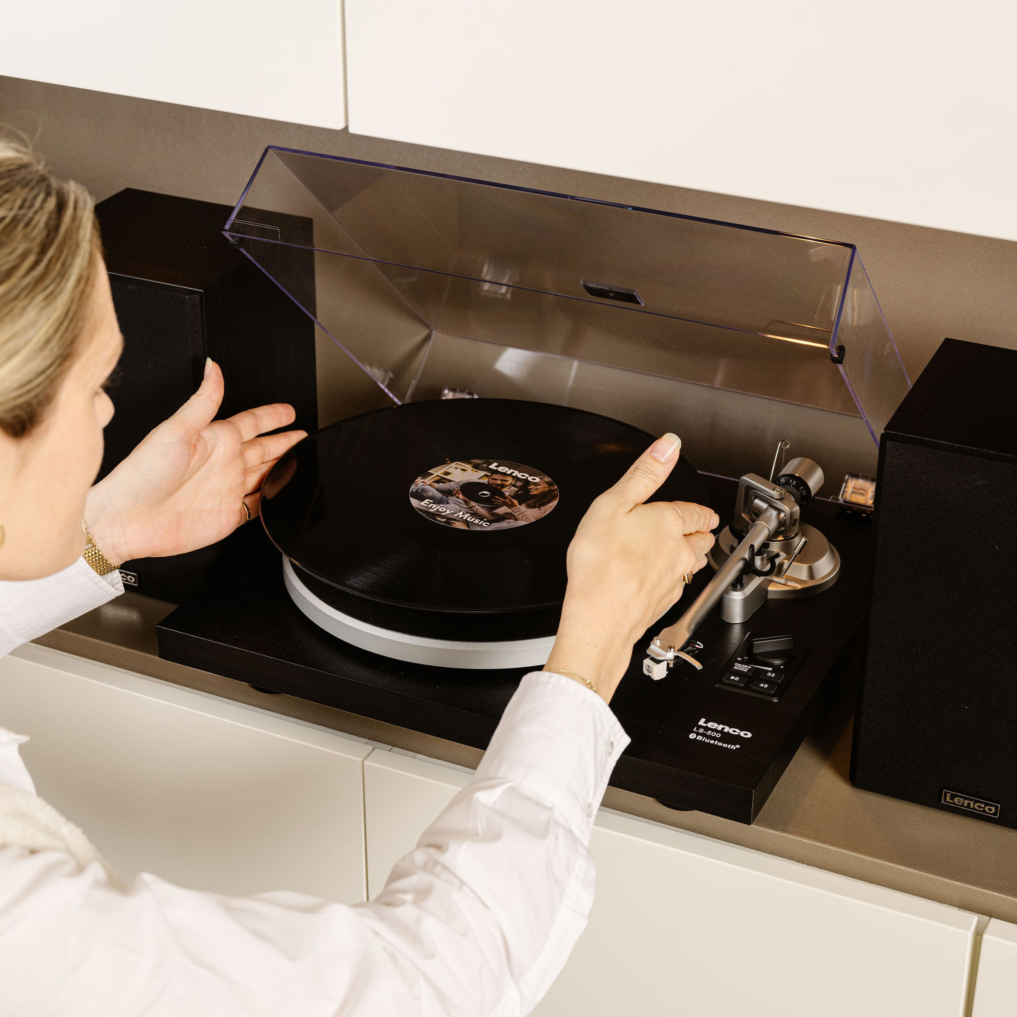 Kobieta obsługuje gramofon. Czarna płyta winylowa leży na talerzu. Głośniki flankują odtwarzacz.