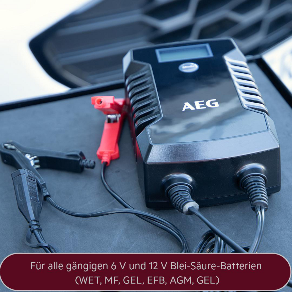Schwarzes und rotes AEG Batterieladegerät mit Kabeln und Klemmen auf schwarzer Oberfläche.