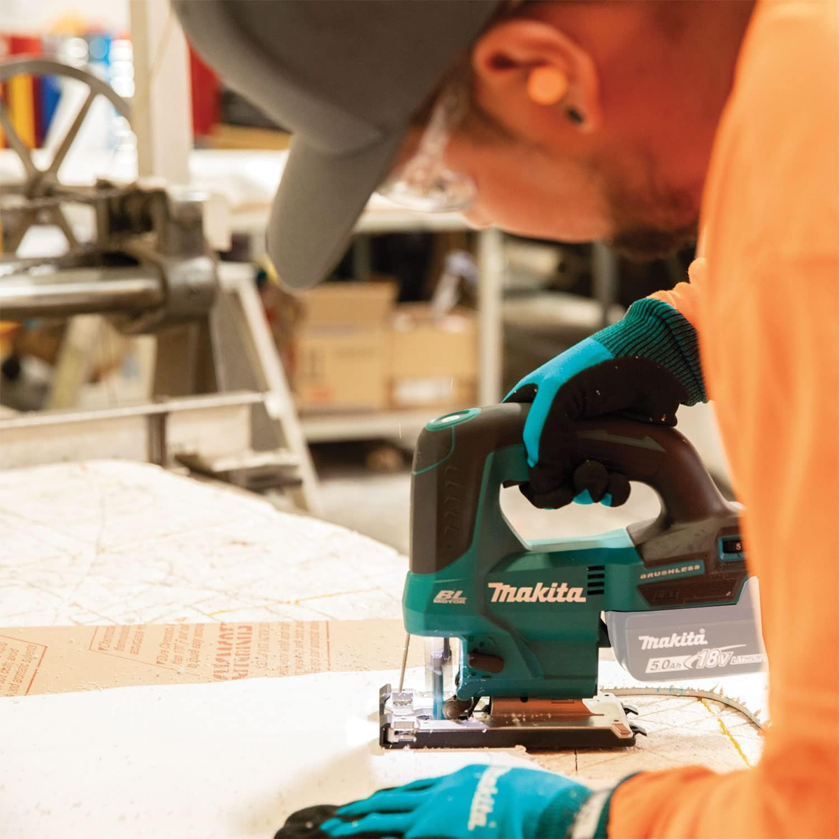 Osoba używająca turkusowo-czarnej wyrzynarki Makita na drewnie, w rękawicach i okularach ochronnych.