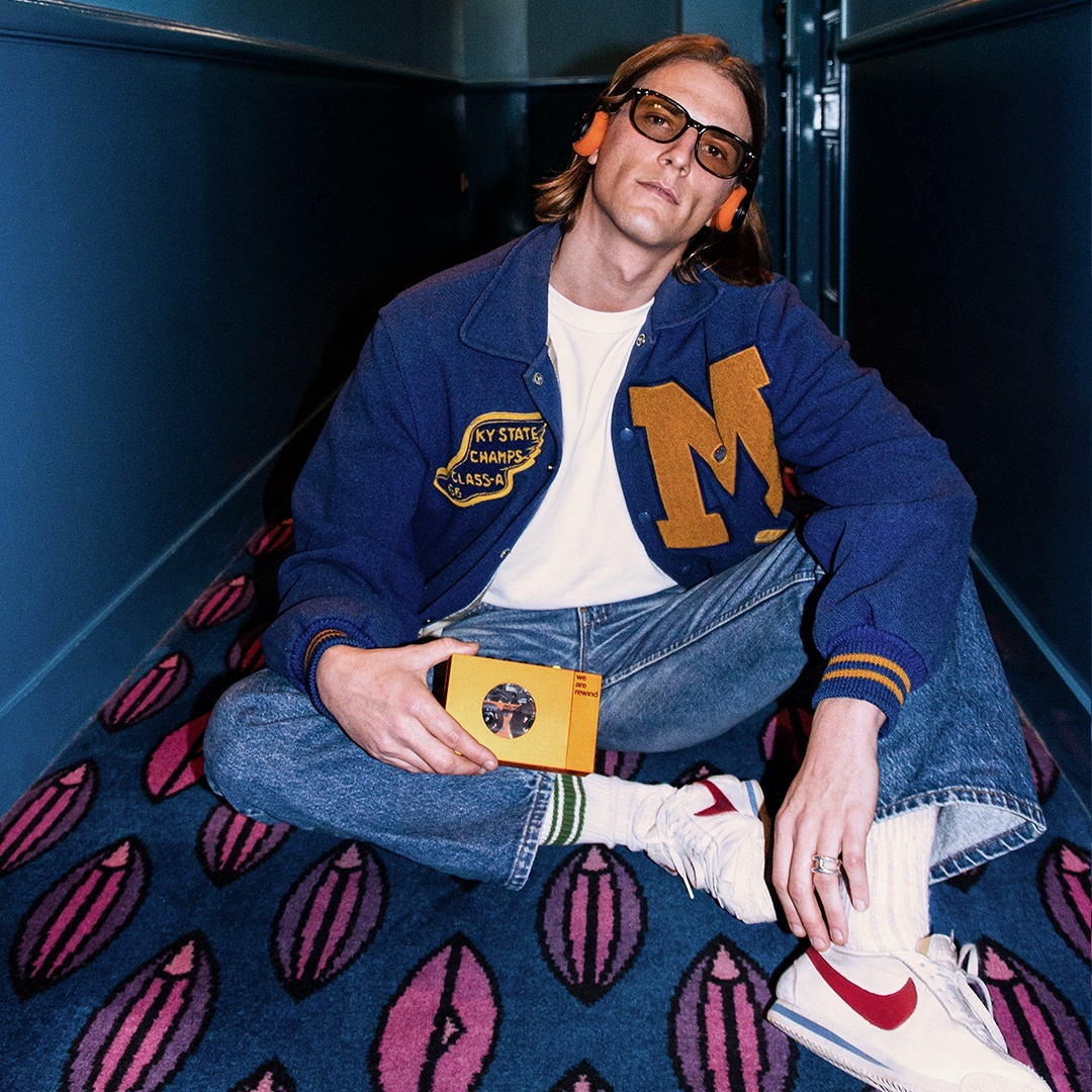 Un homme assis dans un couloir portant des lunettes et un casque, tenant une cassette. Il porte une veste.