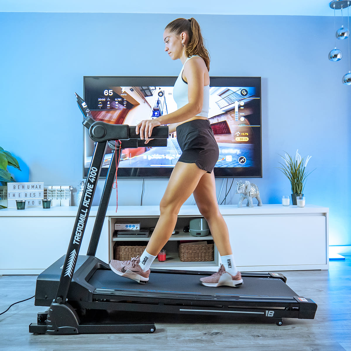Una mujer en una cinta de correr en una habitación con un televisor grande, un gabinete blanco e iluminación azul.