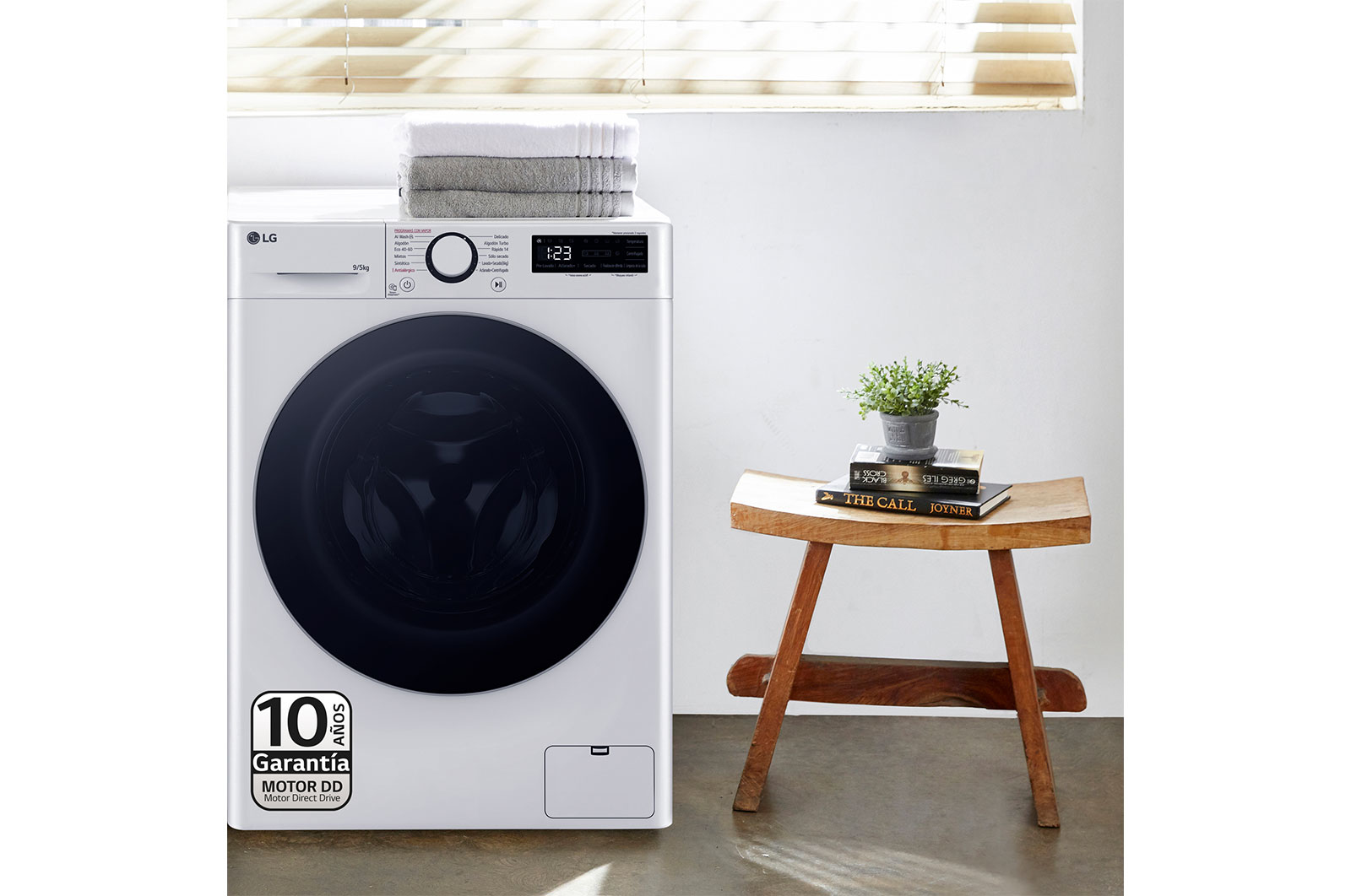 Biała pralka z ręcznikami i małą rośliną na drewnianym stołku.