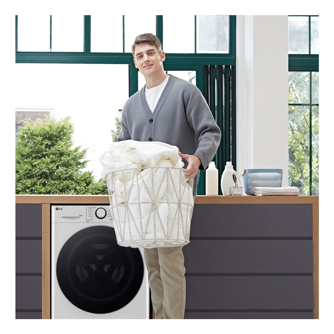 Mężczyzna uśmiecha się, trzymając kosz na pranie wypełniony białymi ubraniami. Pralka i okno w tle.