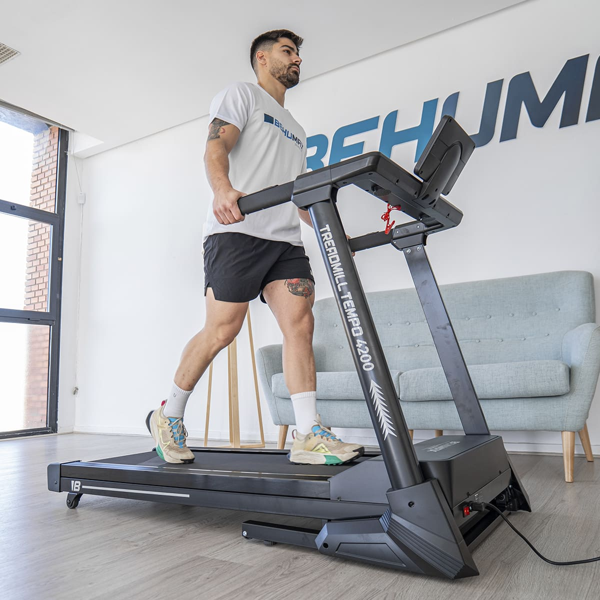 Hombre caminando en una cinta de correr. Pantalones cortos negros, camiseta blanca y zapatillas. Gimnasio.