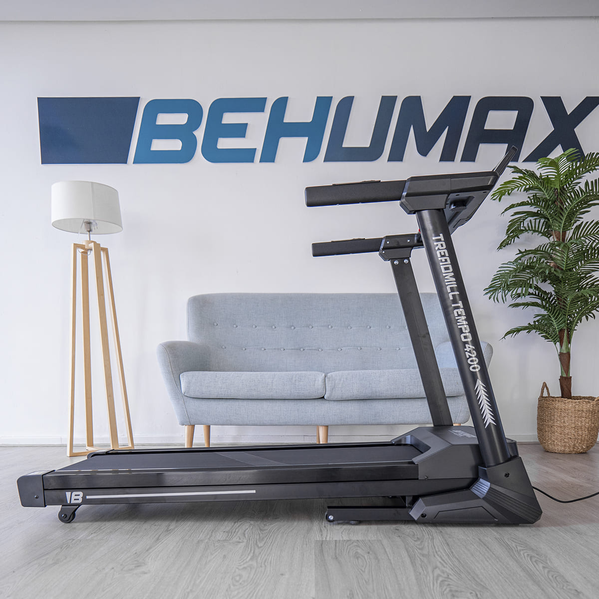 Una cinta de correr negra está en una habitación con un sofá gris, una lámpara y la palabra BEHUMAX en la pared.