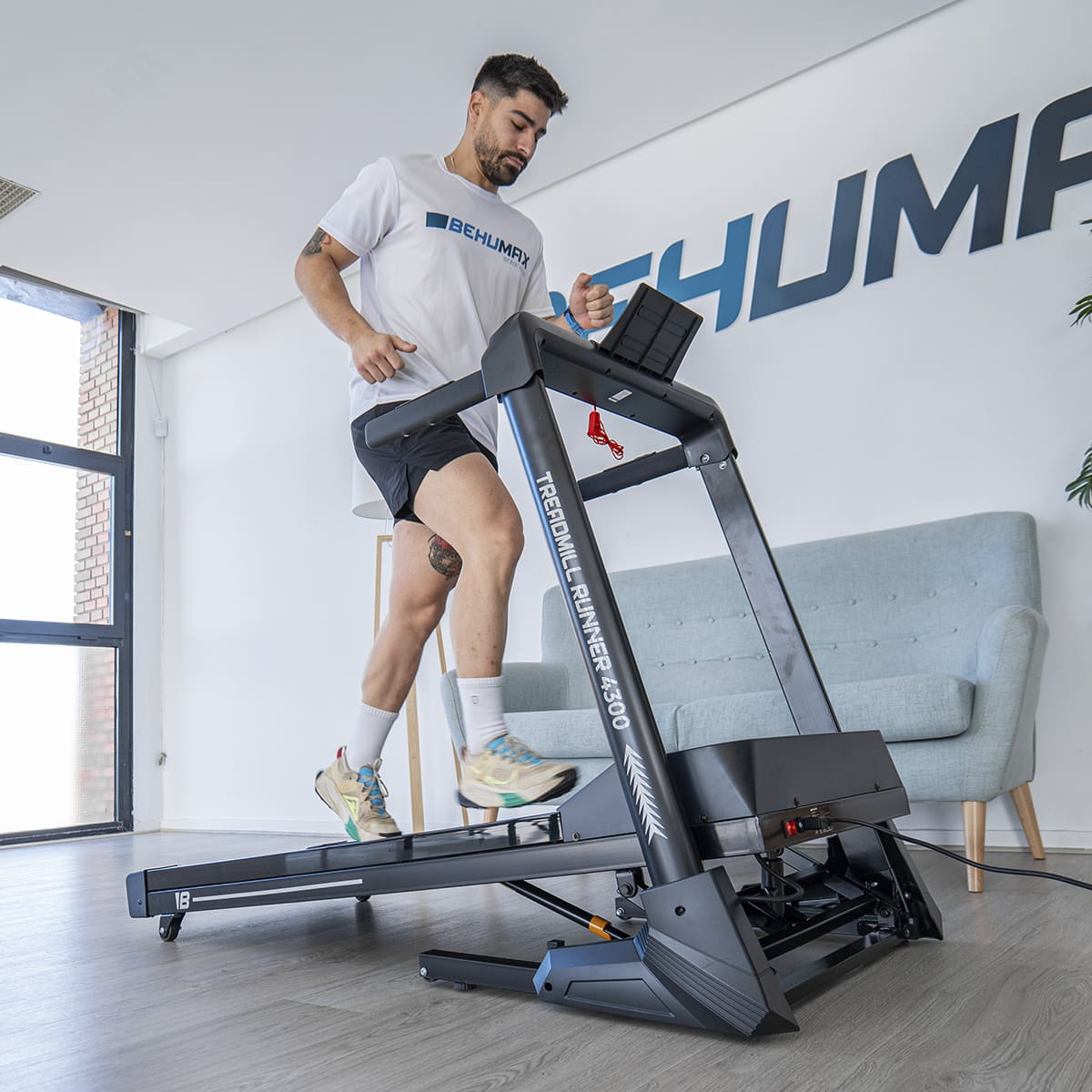 Un hombre corre en una cinta de correr. Un sofá está en el fondo. La cinta lee "Treadmill Runner 4300".