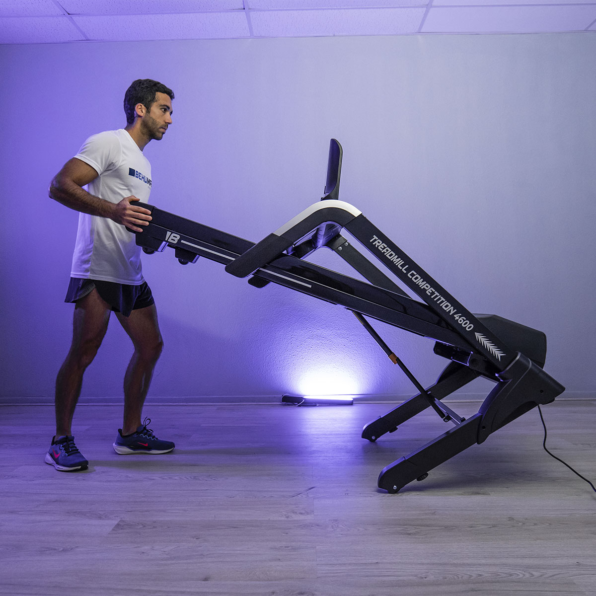 Hombre plegando una cinta de correr. Camiseta blanca, pantalones cortos negros, en una habitación con iluminación morada.