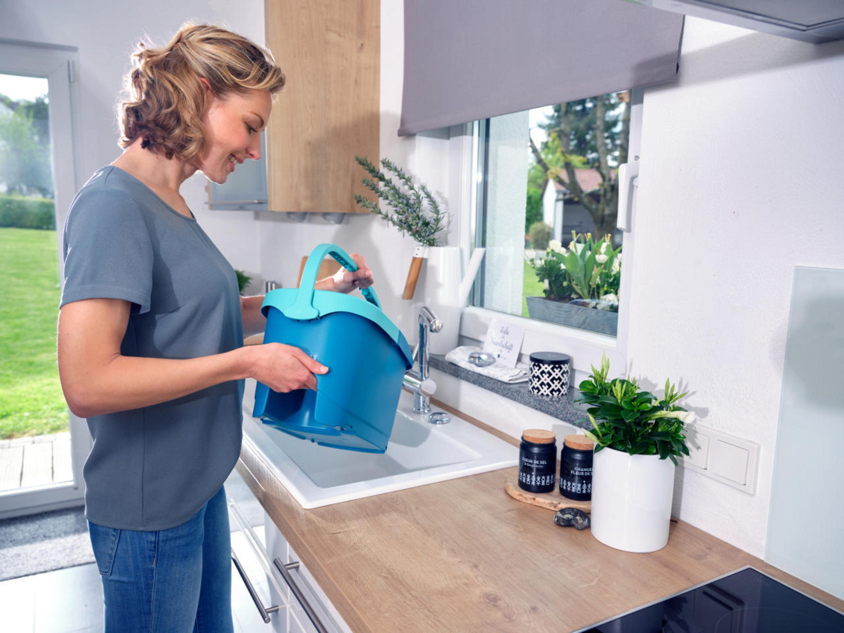 Kobieta w kuchni trzymająca niebieskie wiadro w pobliżu zlewu i okna.