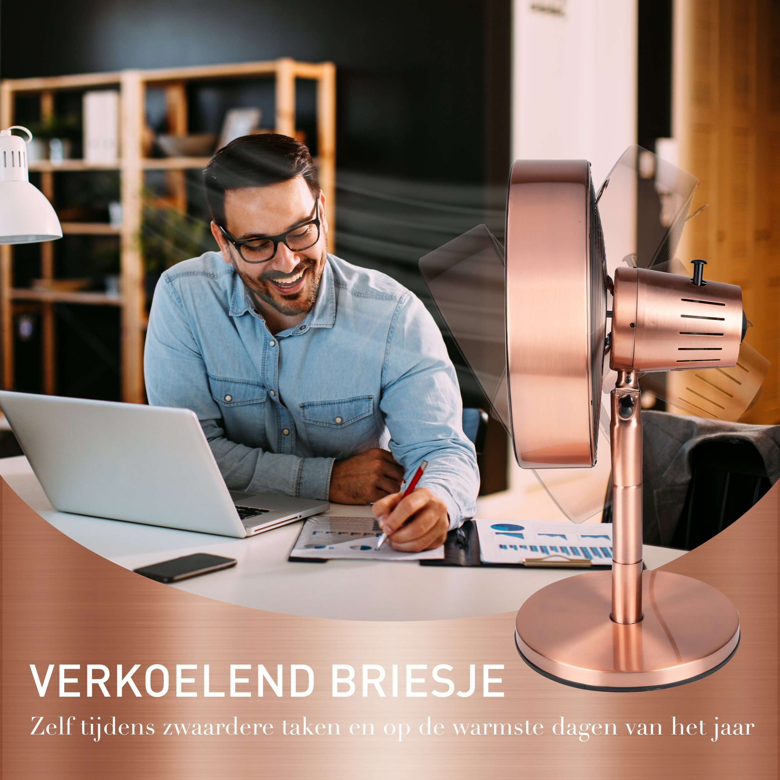 Un homme portant des lunettes sourit en travaillant à un bureau avec un ventilateur. Le ventilateur est cuivré.