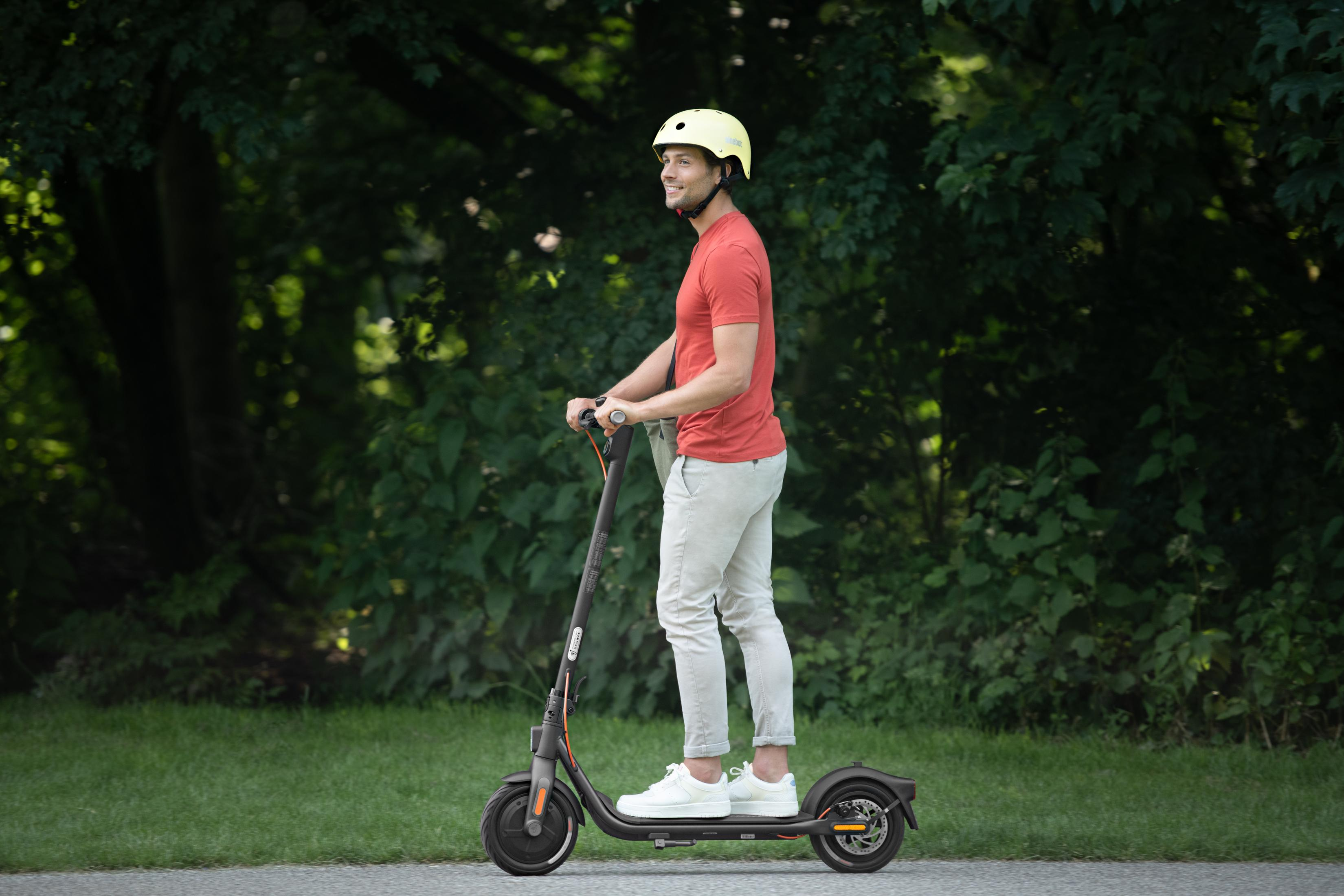 Ein Mann fährt mit einem Elektroroller auf einem Weg in einem Park. Er trägt einen Helm und ein rotes Shirt.