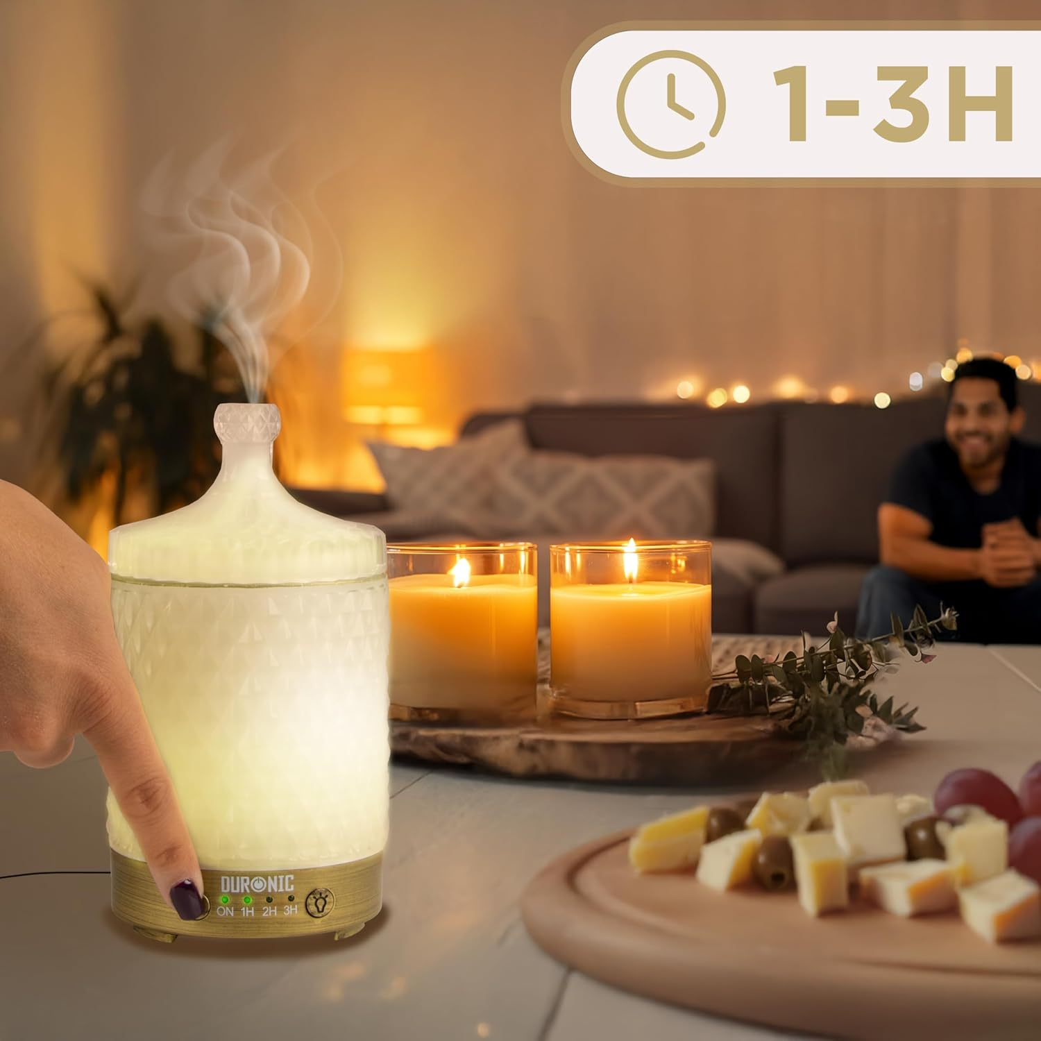 Ein beleuchteter Aromadiffusor, zu dem eine Hand greift. Zwei Kerzen und eine Essensplatte im Hintergrund.