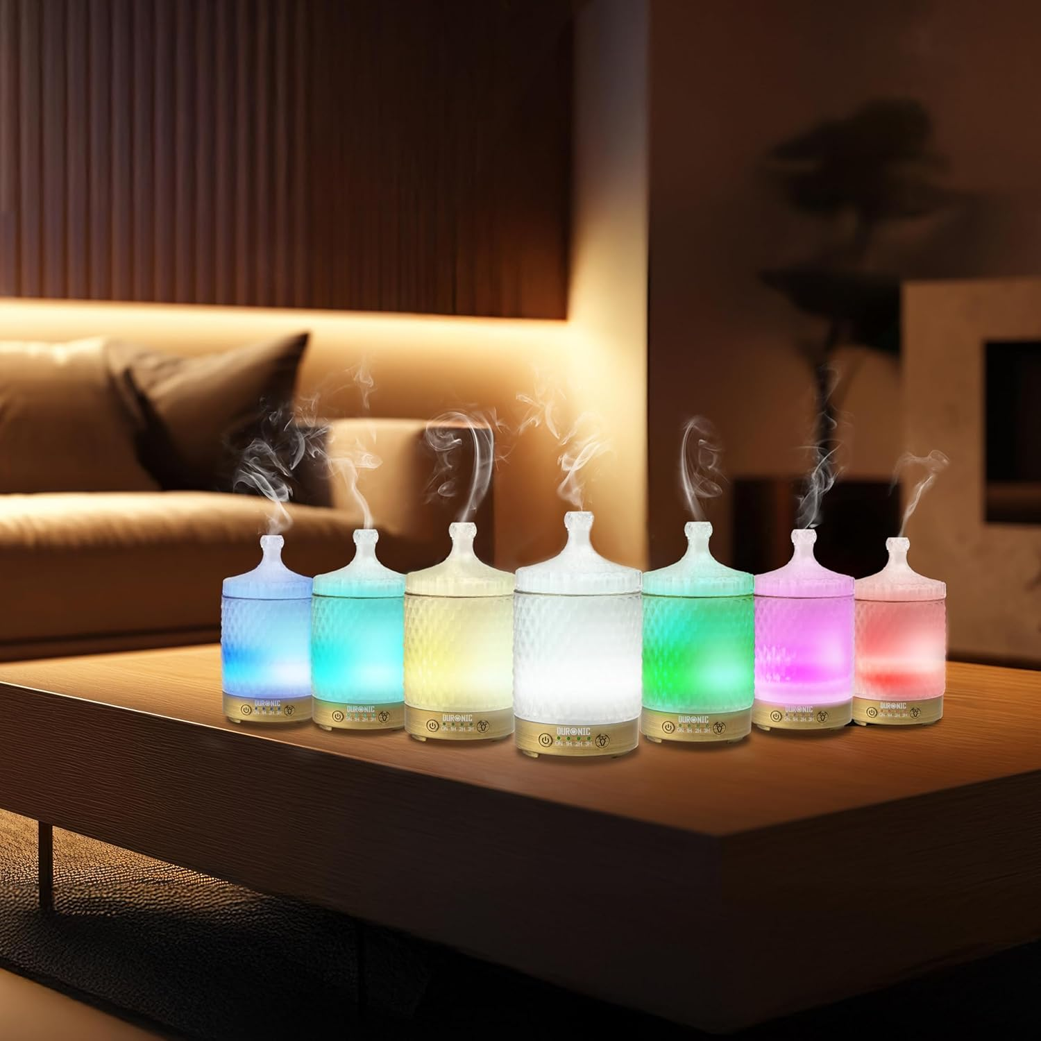 Sieben beleuchtete, farbenfrohe Aromatherapie-Diffusoren auf einem Holztisch, im Hintergrund eine Couch.