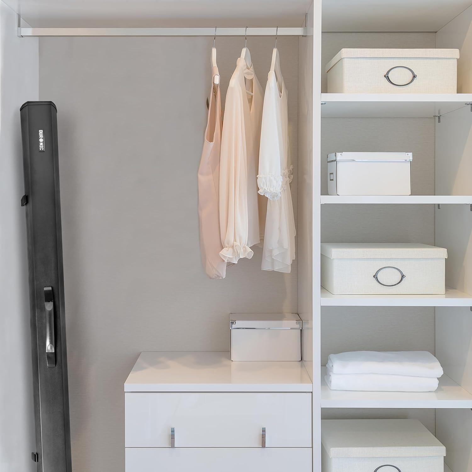 Ein weißer Schrank mit Kleidung darin, weißen Boxen auf Regalen und einem schwarzen Koffer links.