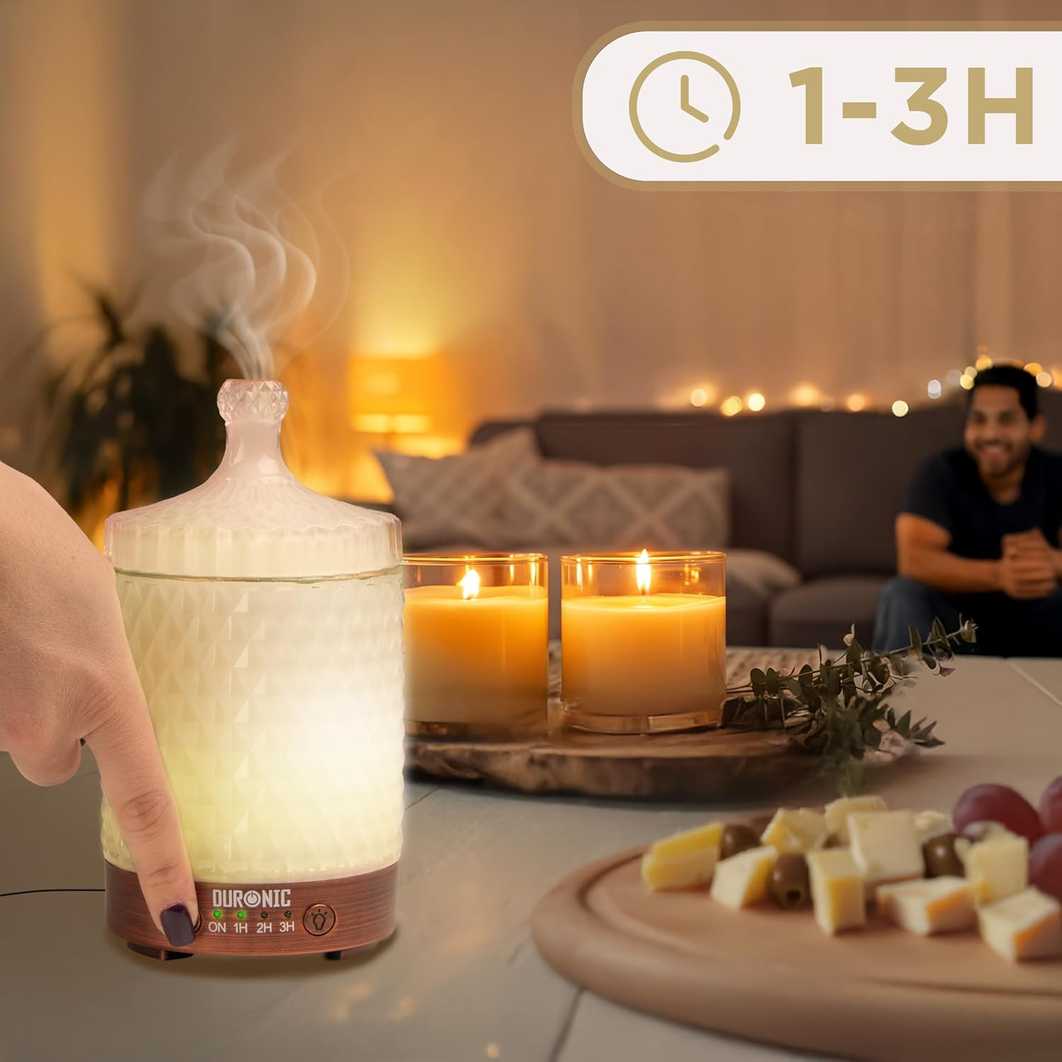 Ein beleuchteter ätherischer Öldiffusor, wobei eine Hand einen Knopf drückt. Auf dem Tisch stehen zwei Kerzen und Snacks.