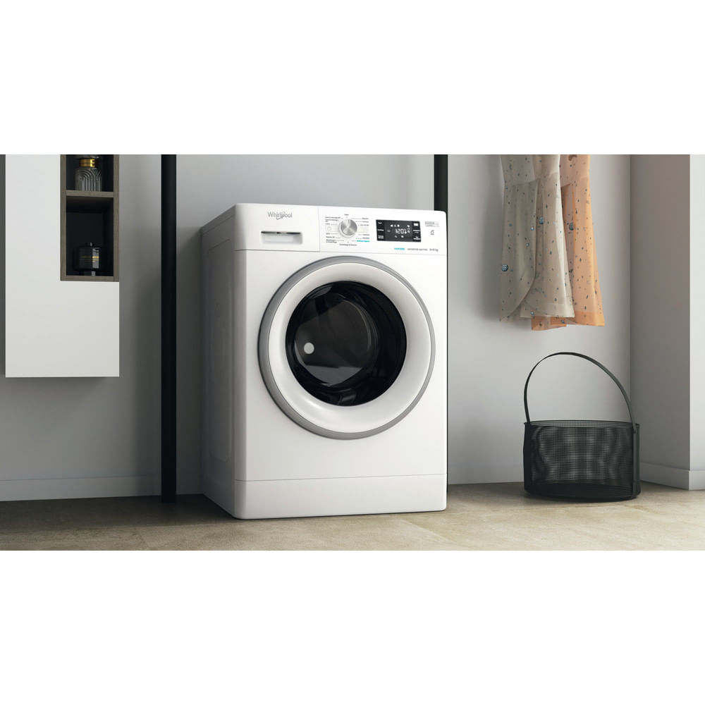 Una lavatrice bianca Whirlpool. Un cestino con manico nero e vestiti appesi nelle vicinanze in una stanza.