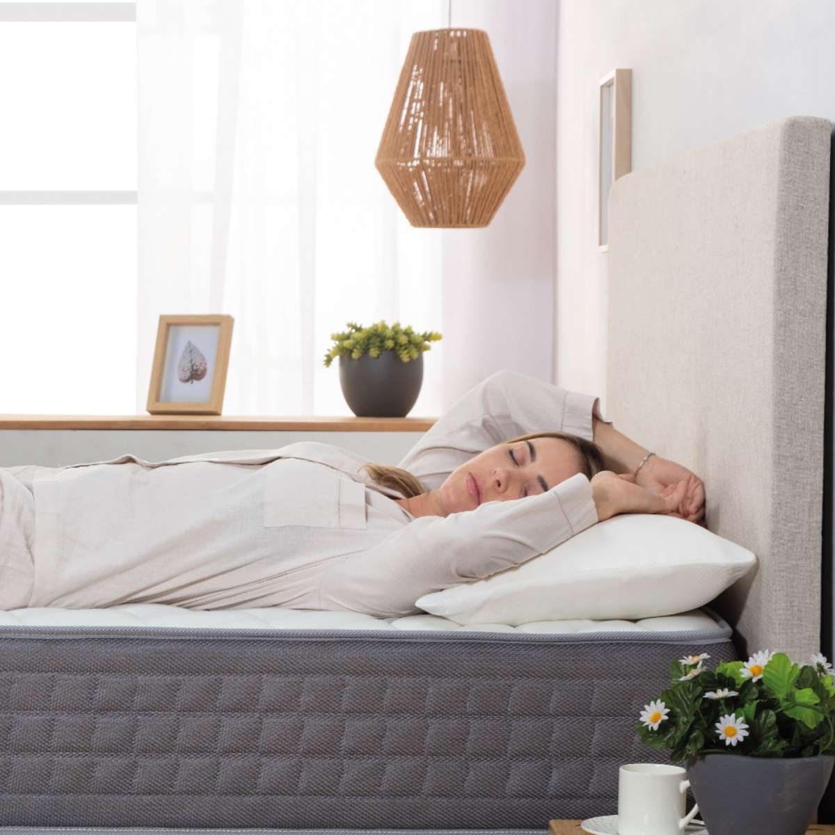 Una mujer en pijama yace en un colchón, con almohada y flores cerca de una ventana.