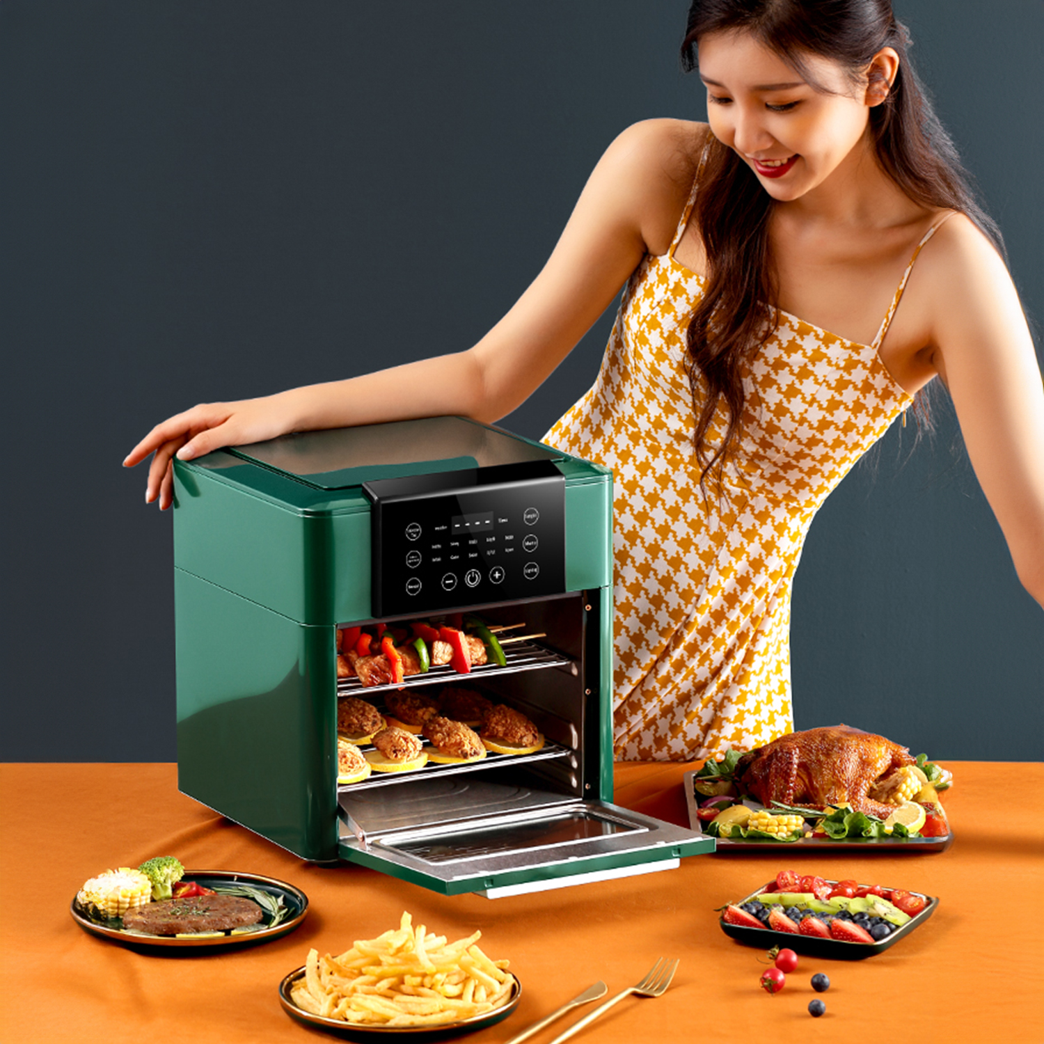 Una mujer sonríe a una freidora de aire con comida en la mesa: bistec, maíz, pollo, papas fritas y brochetas.