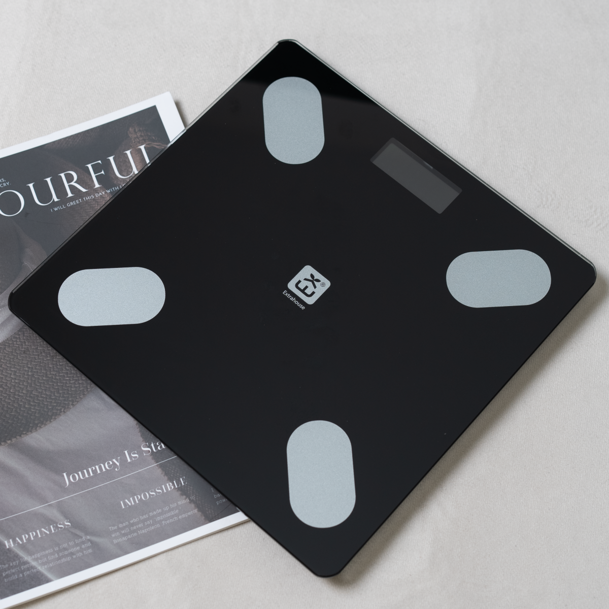 Báscula digital negra con cuatro almohadillas grises sobre superficie blanca, revista detrás.