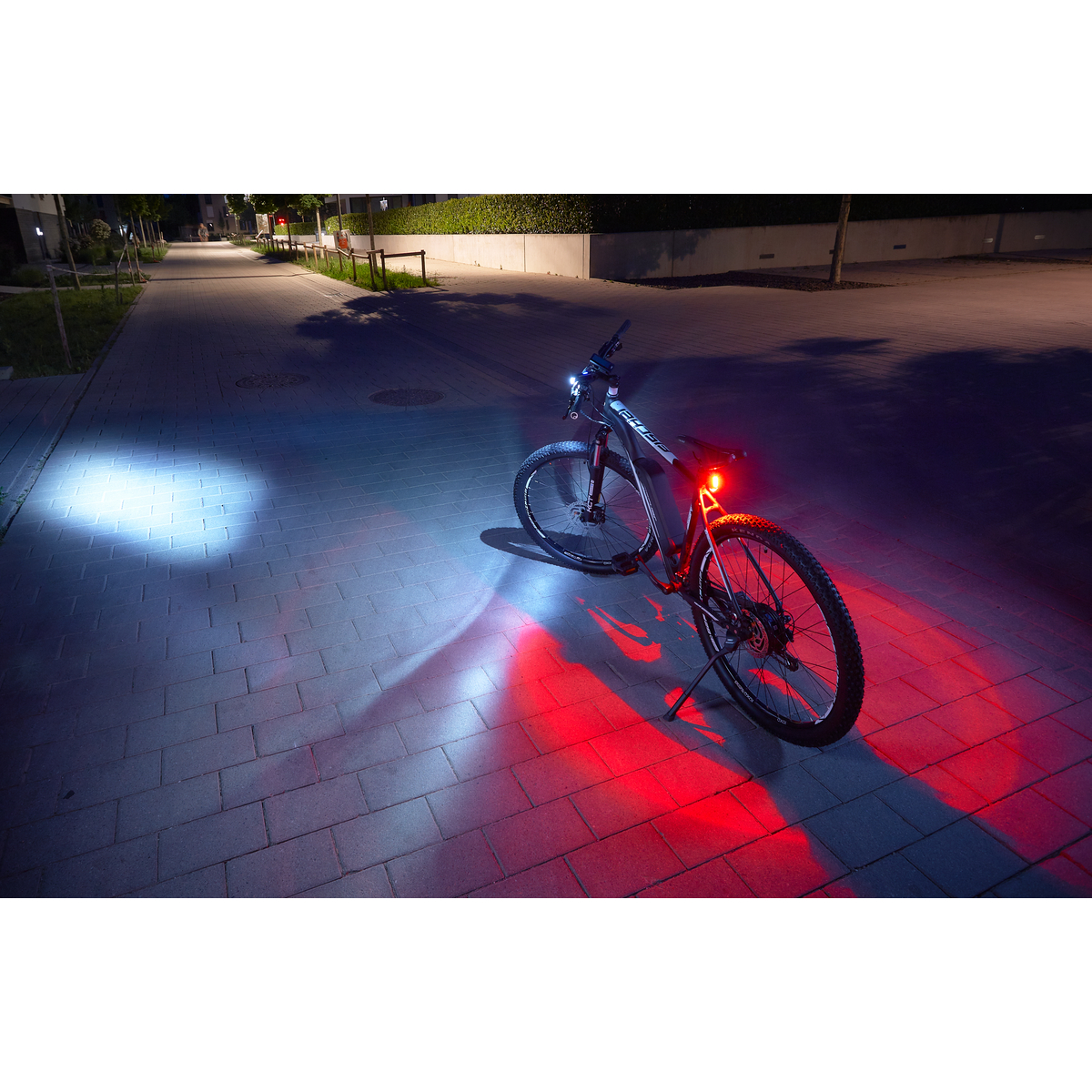 Ein schwarzes Fahrrad mit roten und weißen Lichtern steht nachts auf einer Backsteinstraße, im Hintergrund Straßenlaternen.