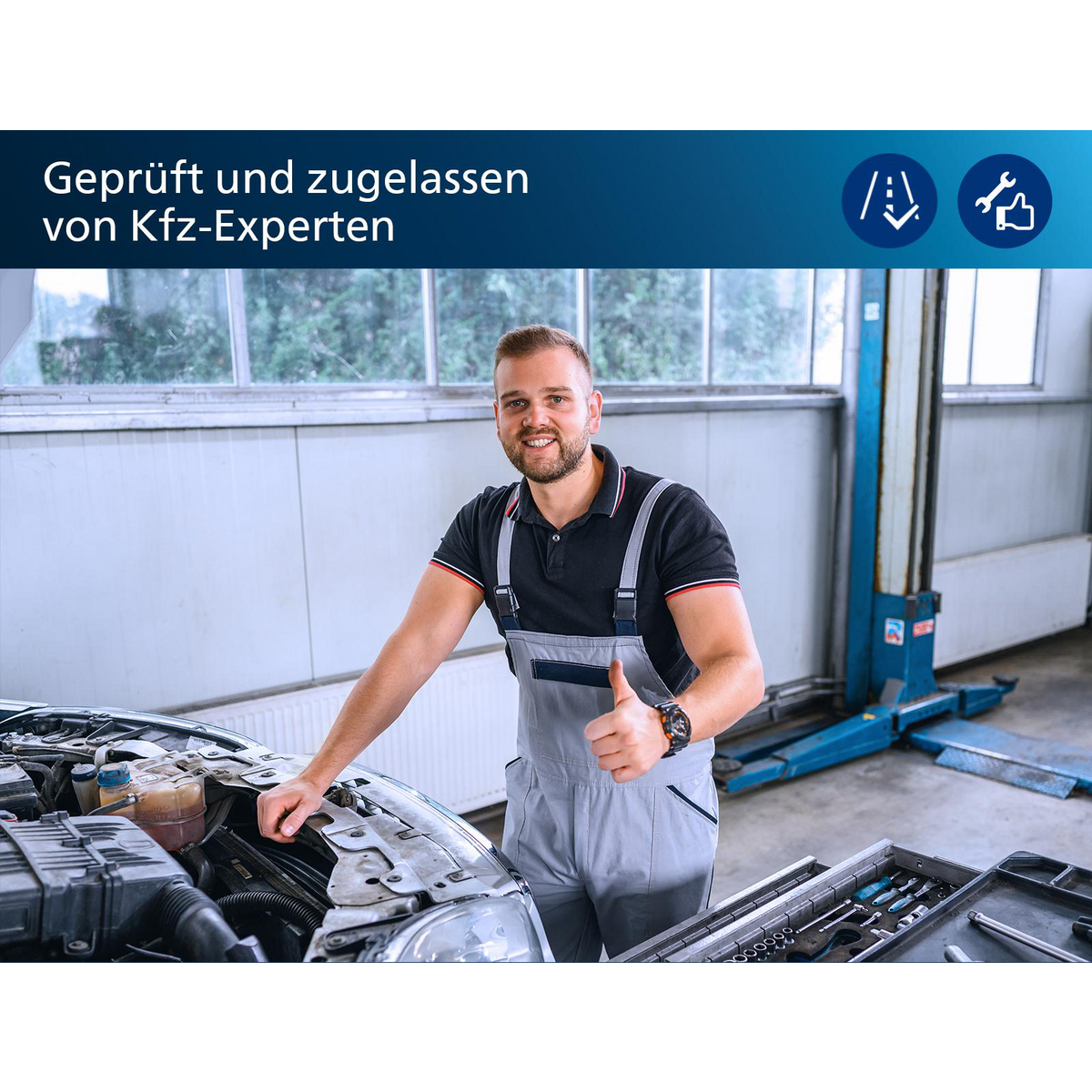 Mechaniker im Overall gibt Daumen hoch, lächelt in die Kamera. Auto-Motor offen im Hintergrund, Werkzeuge sichtbar.