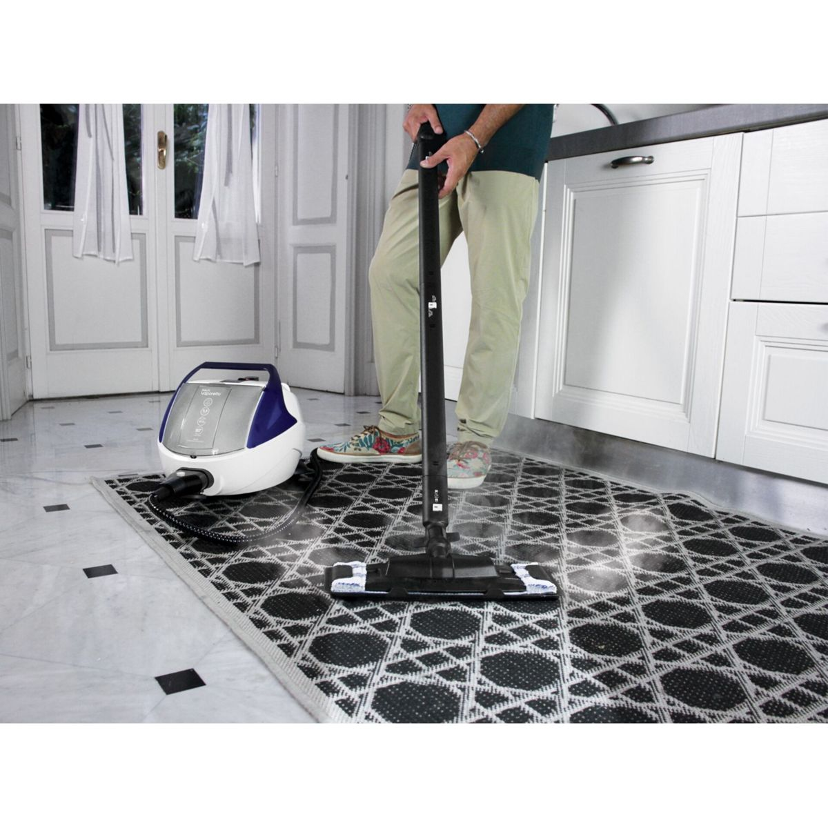 Osoba używa parownicy na wzorzystym dywanie w kuchni. Urządzenie jest po lewej stronie.