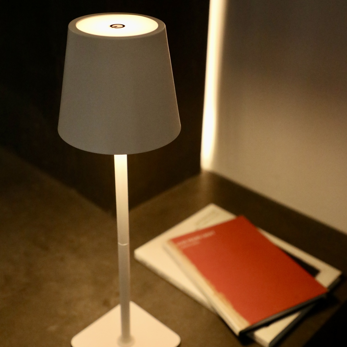 Lámpara de mesa blanca con pantalla blanca y dos libros sobre una mesa con fondo oscuro.