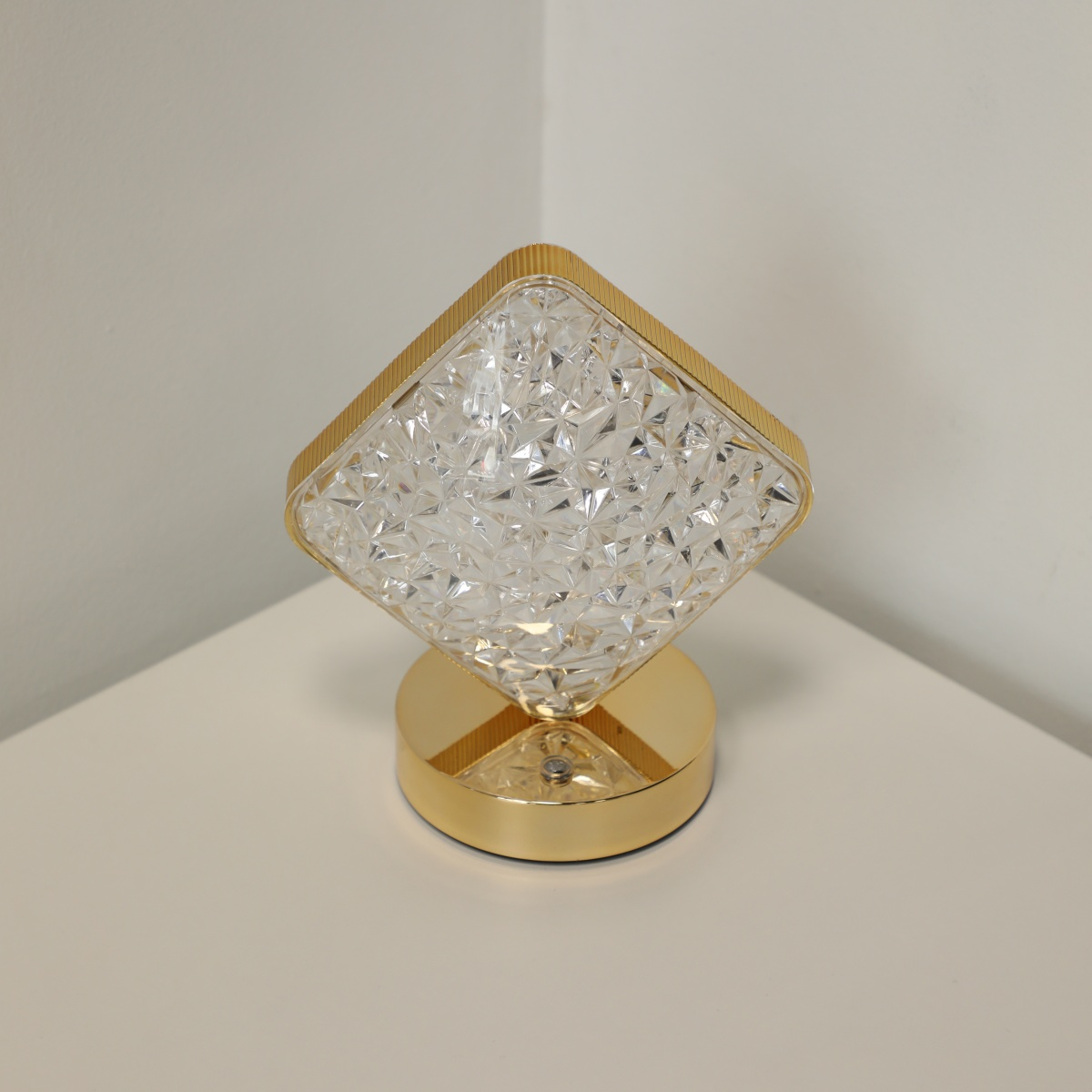 Una lámpara dorada en forma de diamante se encuentra sobre una superficie blanca. Tiene una superficie de vidrio facetada y espejada.