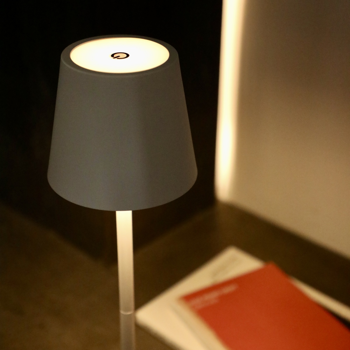 Una lámpara de mesa gris con luz blanca. Está sobre una mesa con un libro rojo y papeles.
