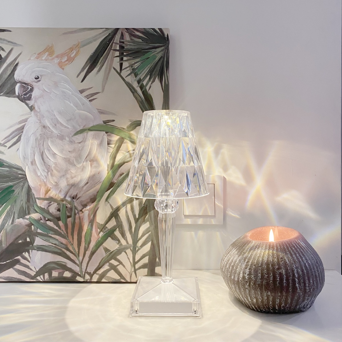 Una lámpara de cristal transparente con una vela encendida. Una pintura de un pájaro blanco detrás.