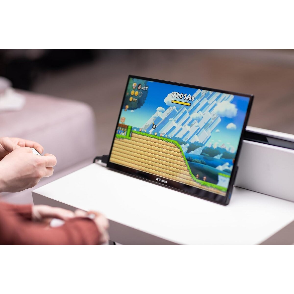 Osoba gra w grę na przenośnym monitorze Verbatim na białym stole, z białą konsolą.