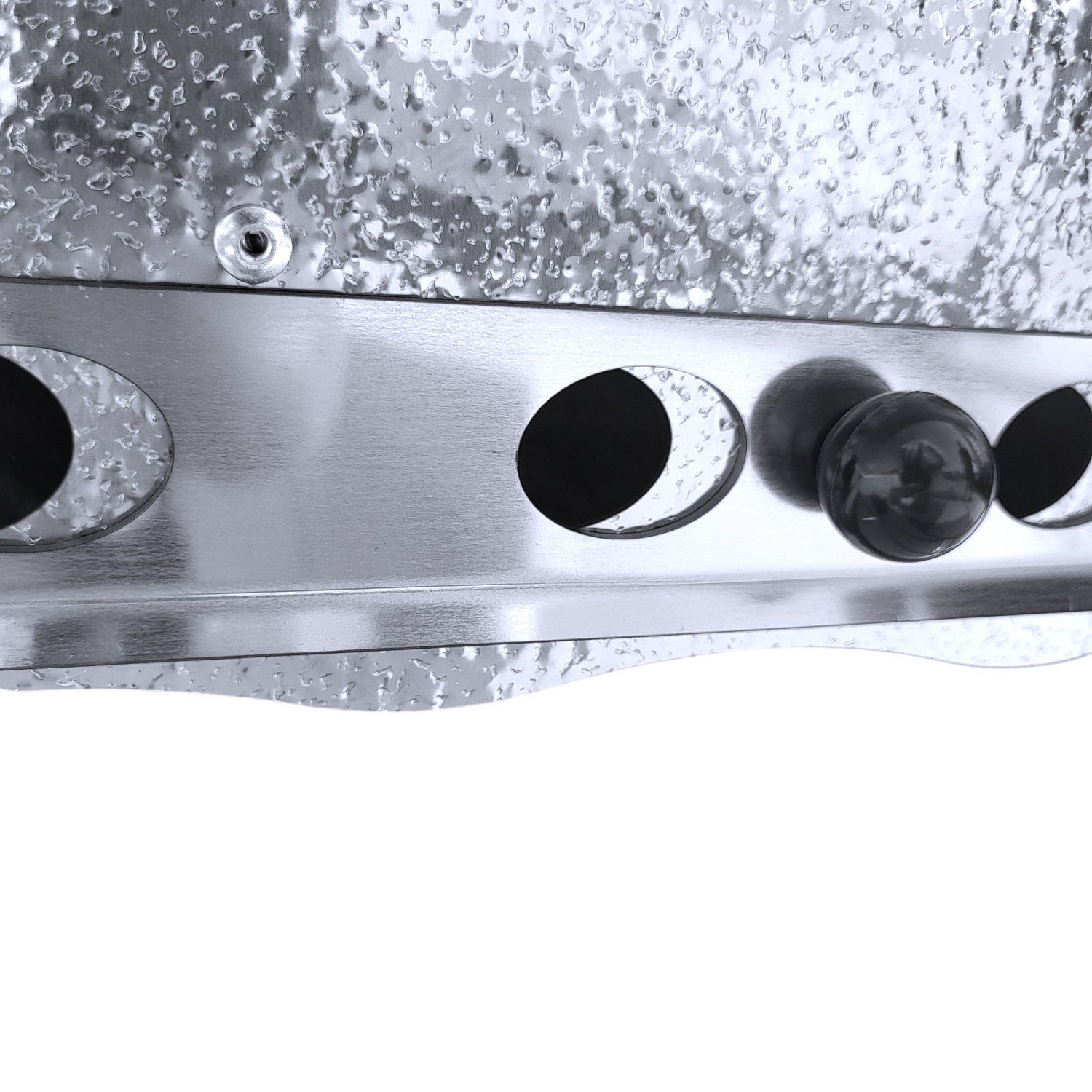 Nahaufnahme einer metallischen Oberfläche mit Löchern und einem schwarzen Knopf. Die Oberfläche ist glänzend und strukturiert.