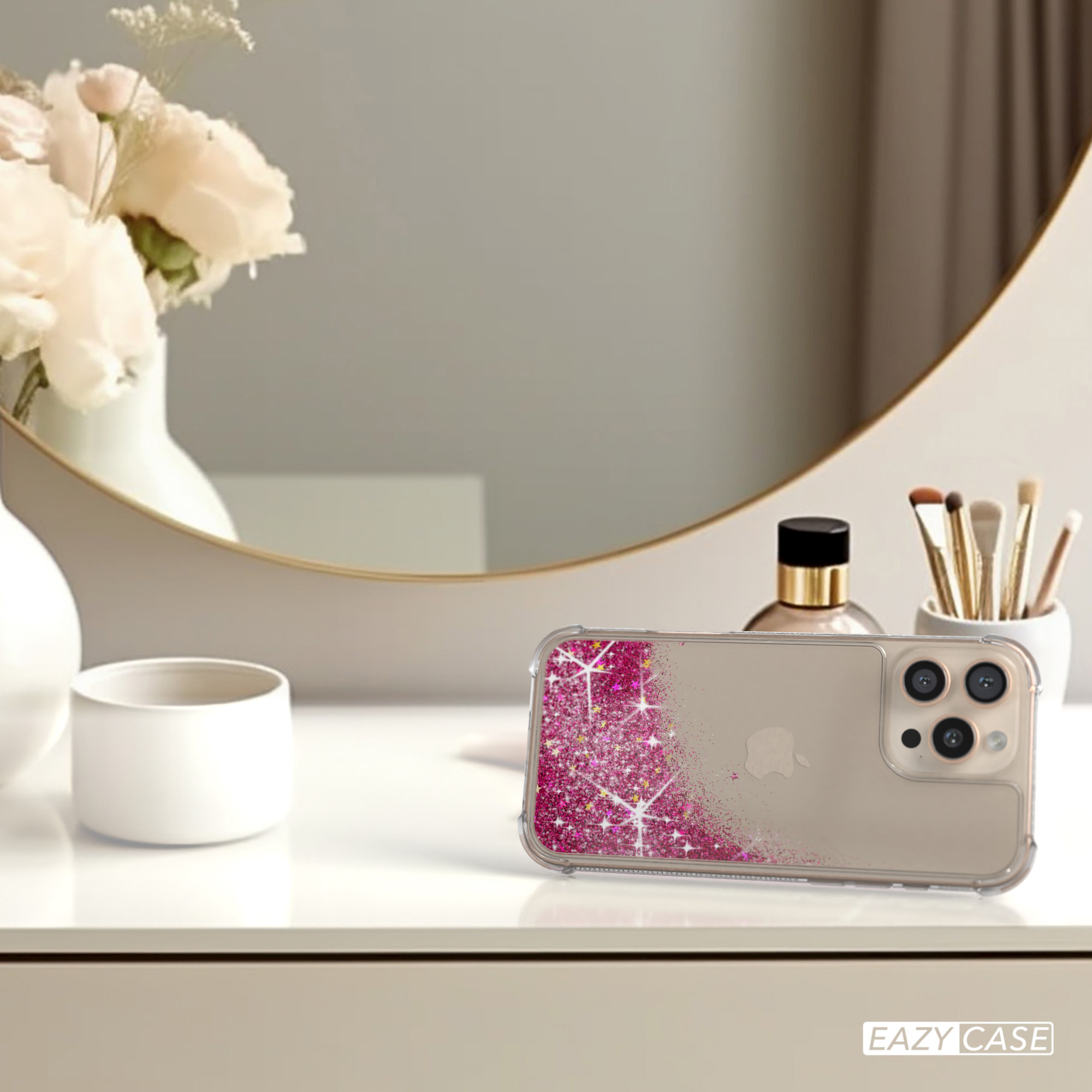 Eine Handyhülle mit pinkfarbenem Glitzer liegt auf einem weißen Tisch mit Spiegel und Blumen im Hintergrund.
