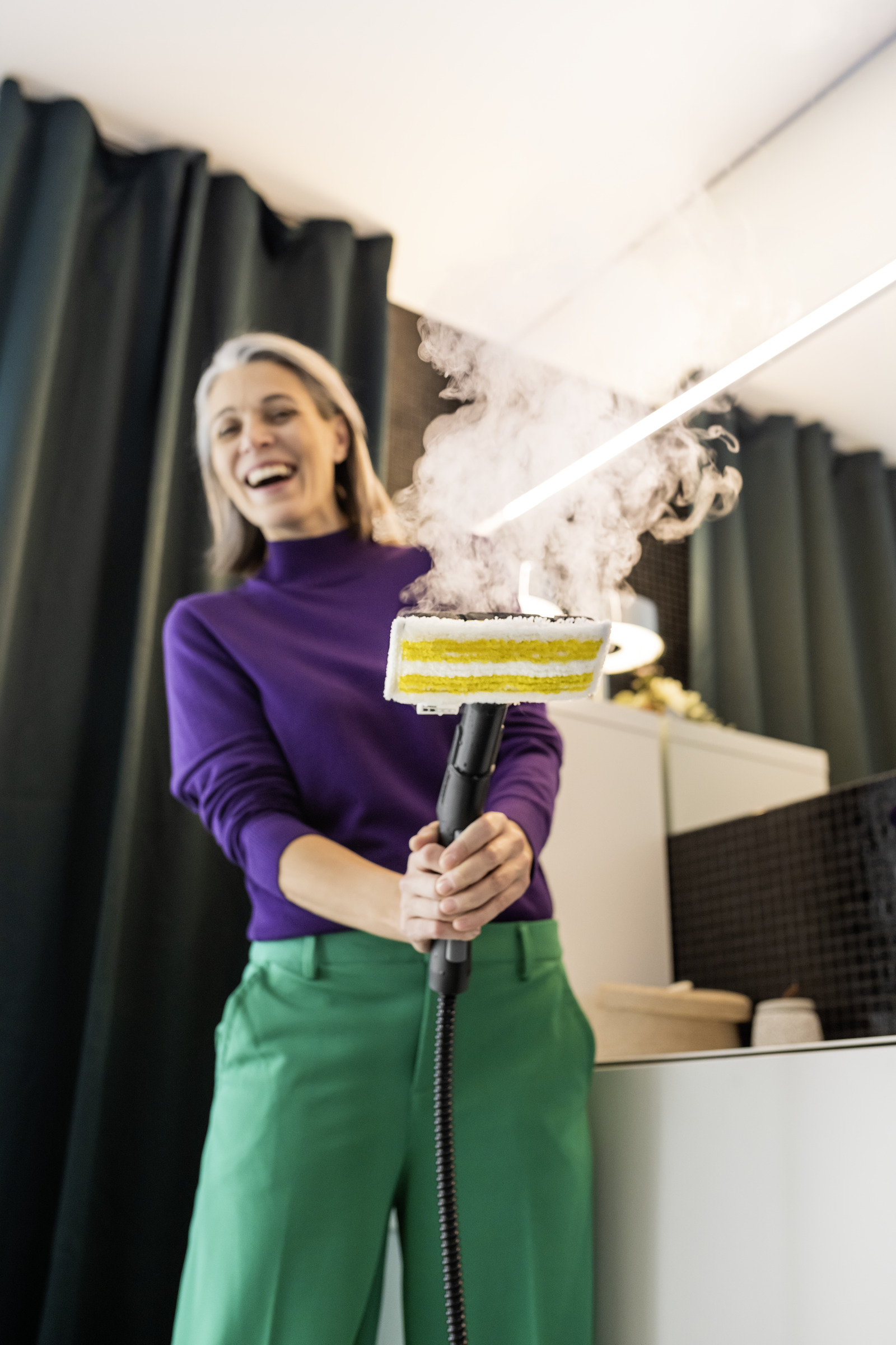 Kobieta śmieje się podczas używania myjki parowej. Wydobywa się para. Ma na sobie fioletową koszulę i zielone spodnie.