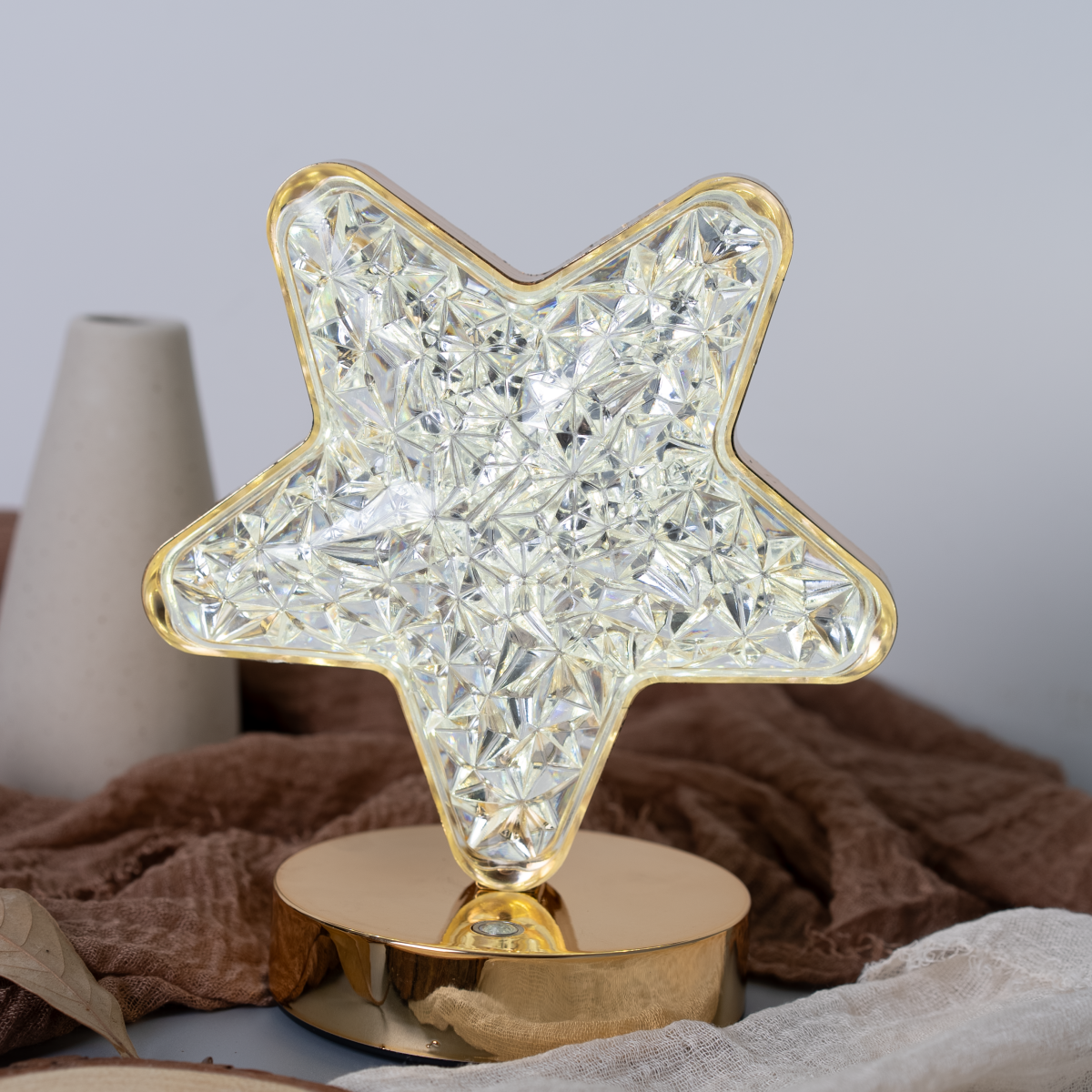 Lámpara dorada en forma de estrella con luz brillante y cristalina. Sobre base dorada. Fondo de tela marrón.