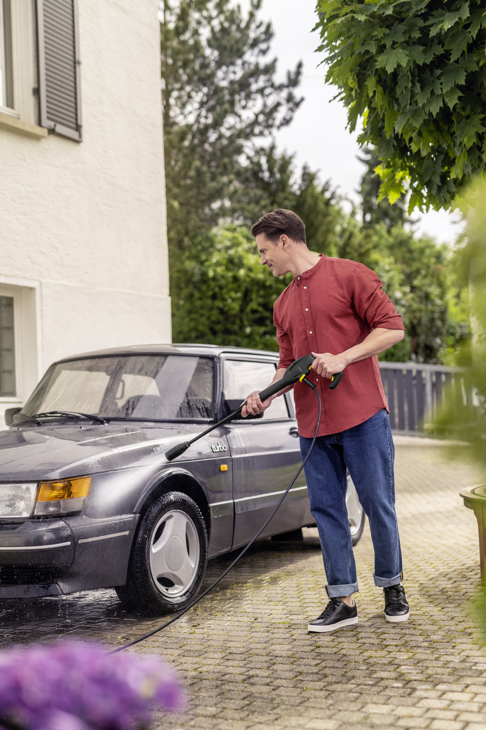 Mann wäscht ein graues Auto mit einem Hochdruckreiniger. Grüne Bäume und ein Haus.