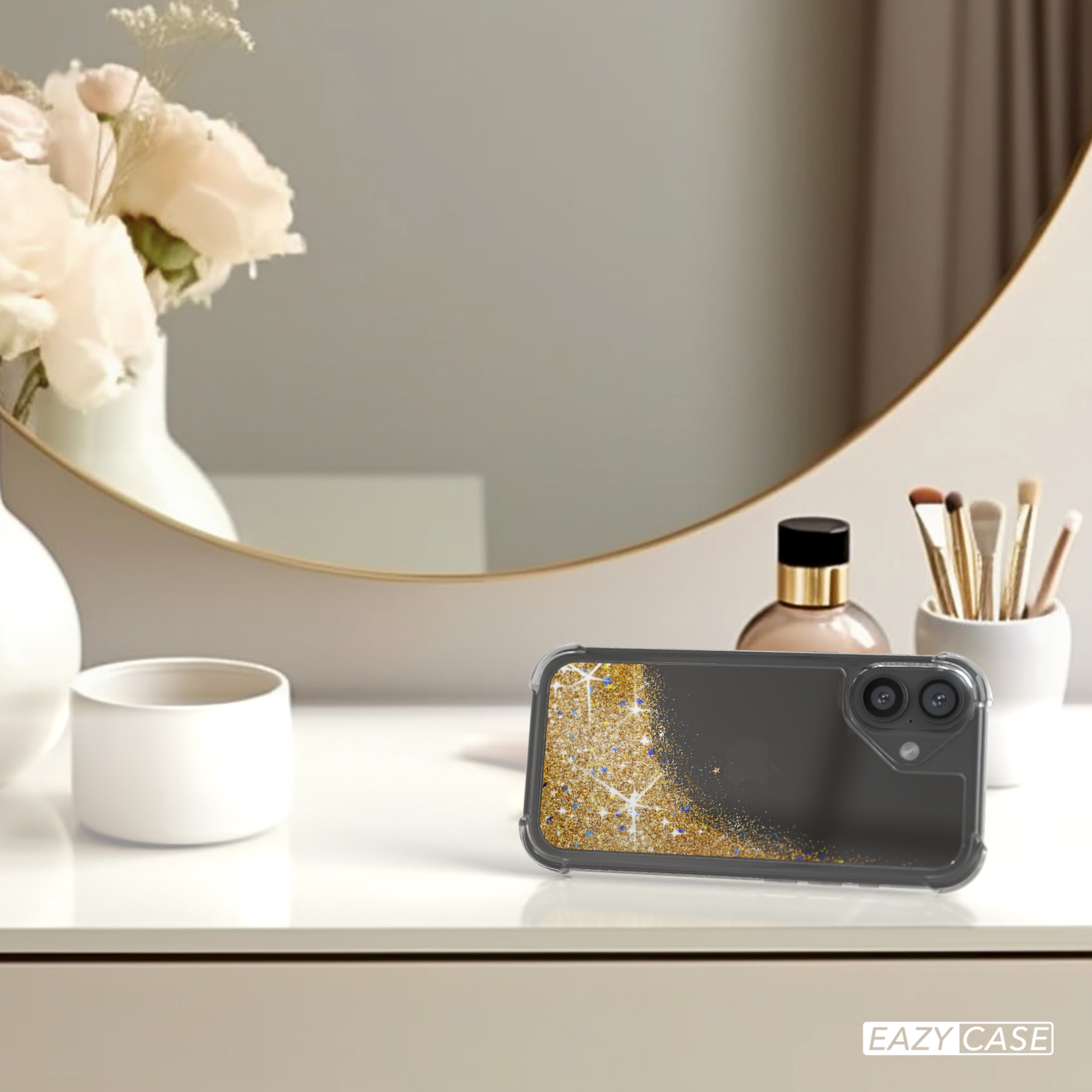 Handyhülle mit goldenem Glitzer auf einem weißen Tisch, in der Nähe eines Spiegels und Blumen.
