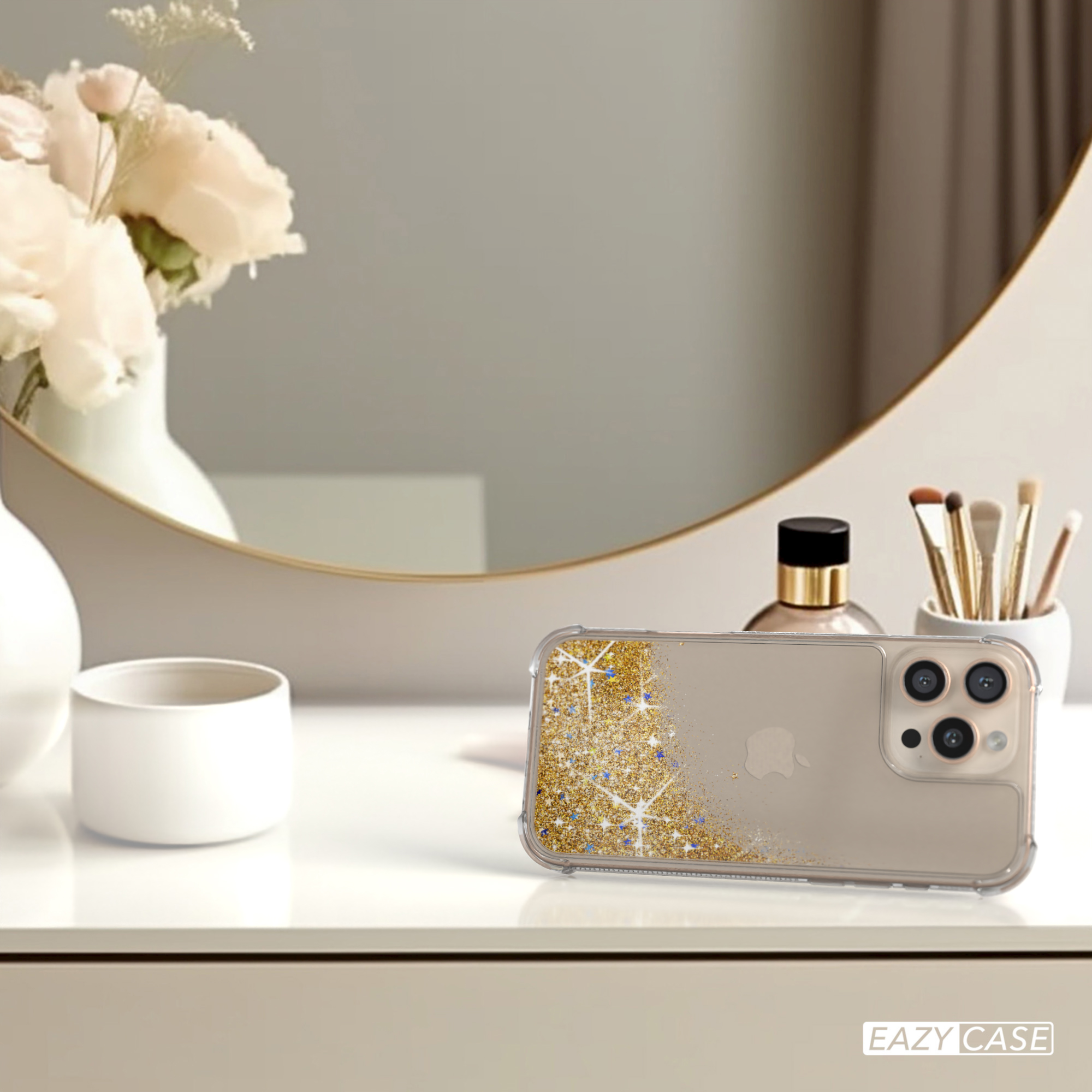 Eine Handyhülle mit goldenem Glitzer auf einem weißen Tisch, in der Nähe eines Spiegels und Blumen. Die Hülle ist klar.