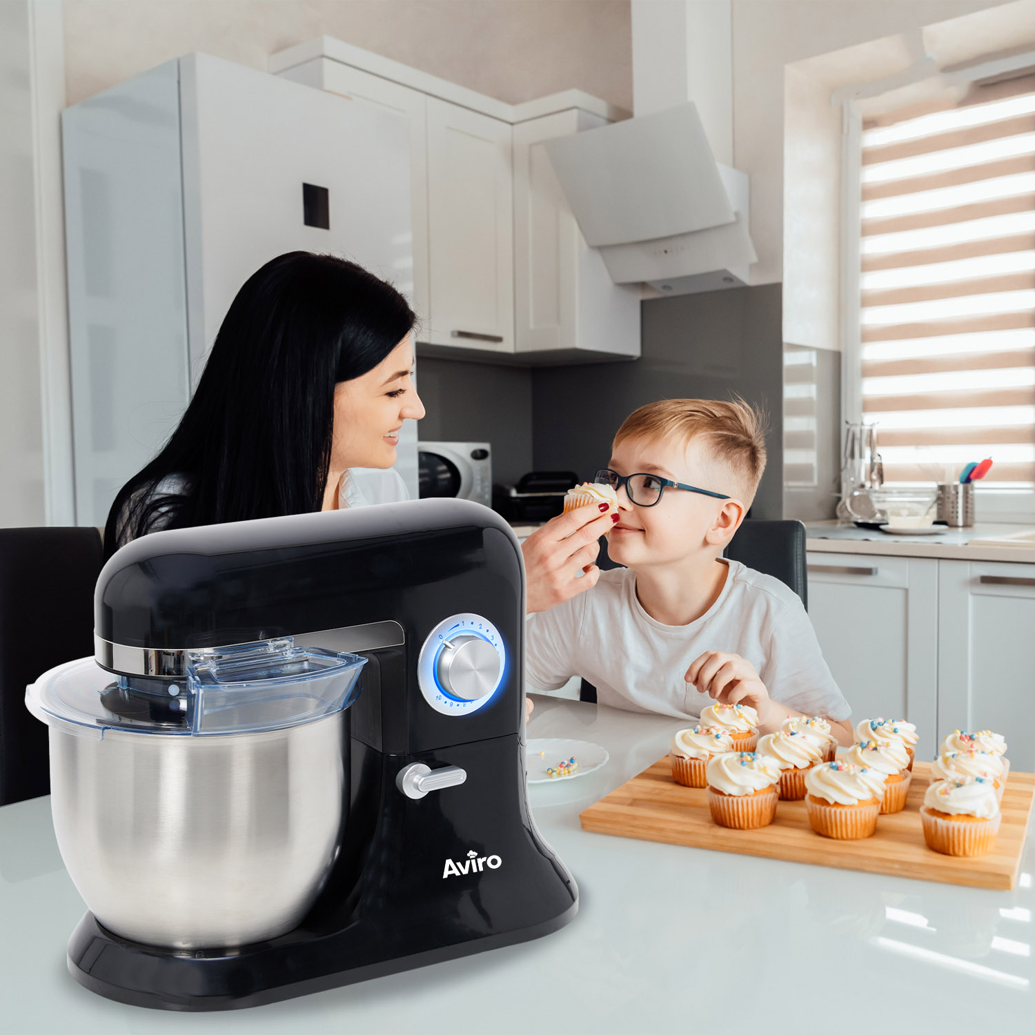 A woman and a boy are in a kitchen. A black mixer is on the table with cupcakes.