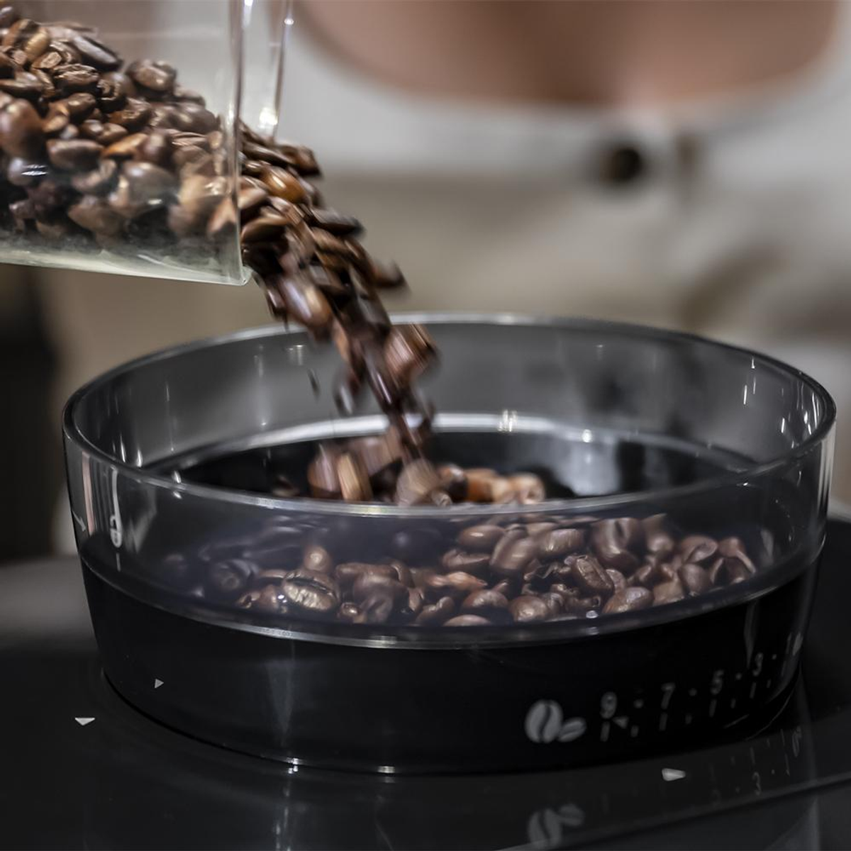 Ziarna kawy wlewane do przezroczystego pojemnika na czarnej powierzchni.