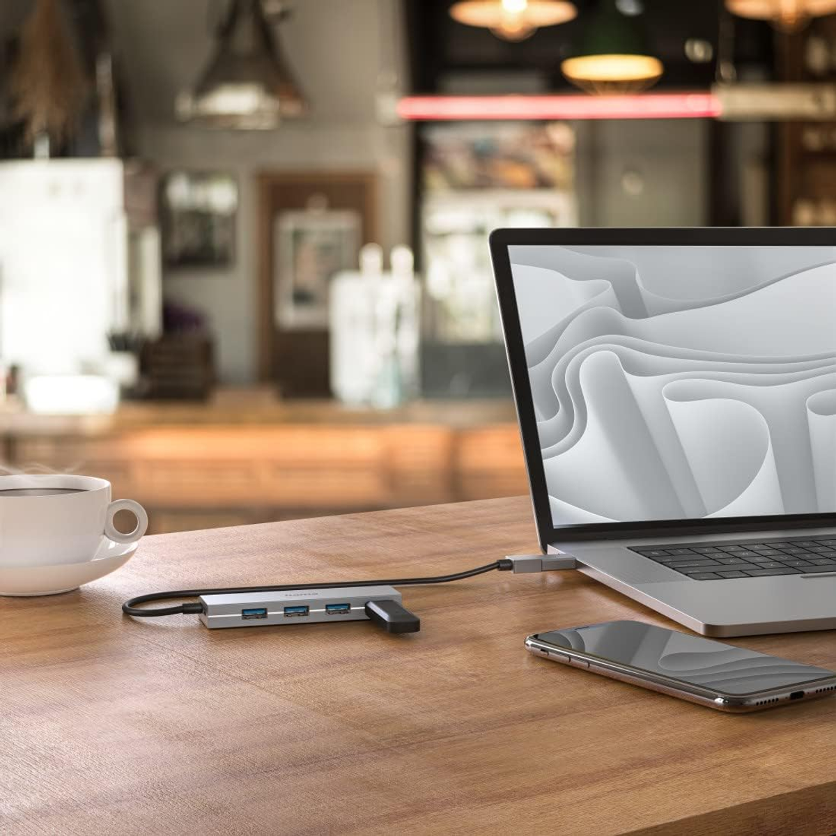 Laptop, hub USB, telefono e tazza su un tavolo di legno. Sfondo sfocato di un caffè.