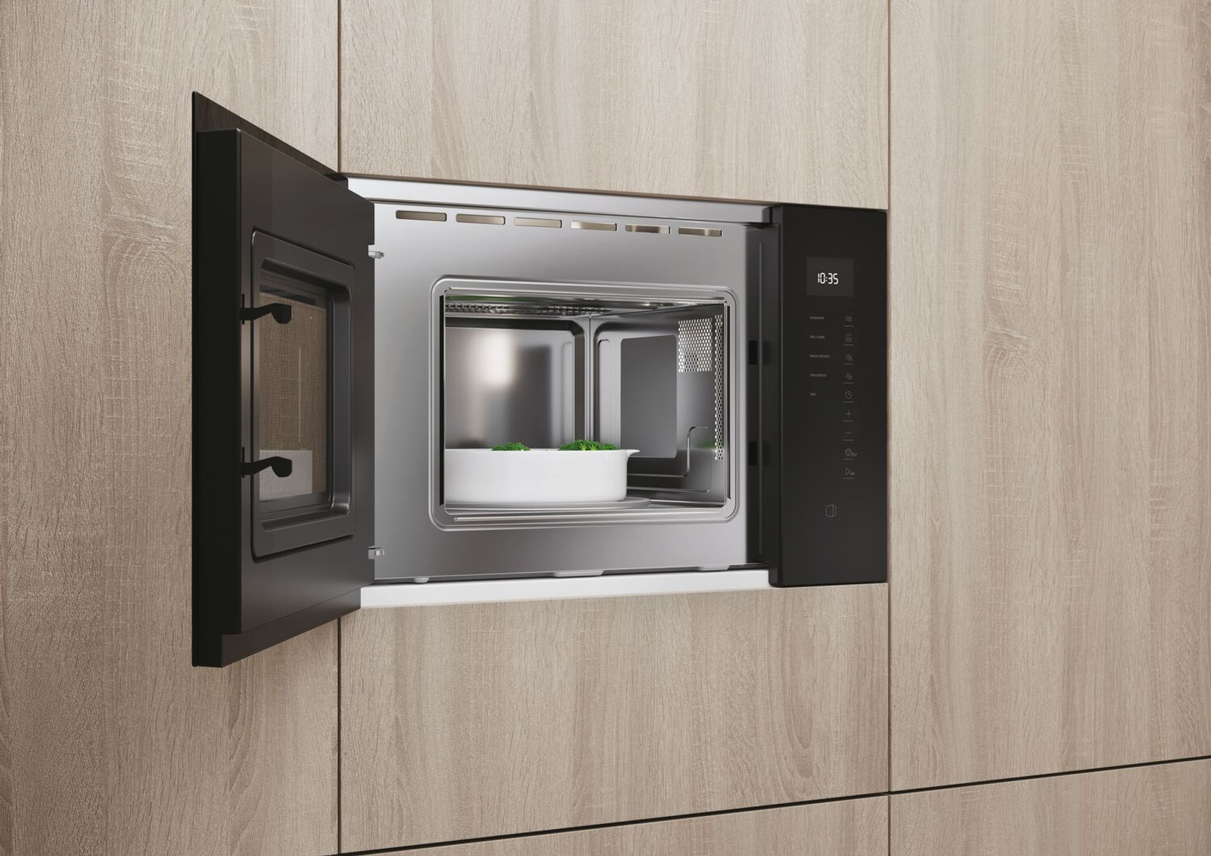 Kuchenka mikrofalowa wbudowana w drewniane szafki, otwarta. Wewnątrz biała miska z zielonym jedzeniem.