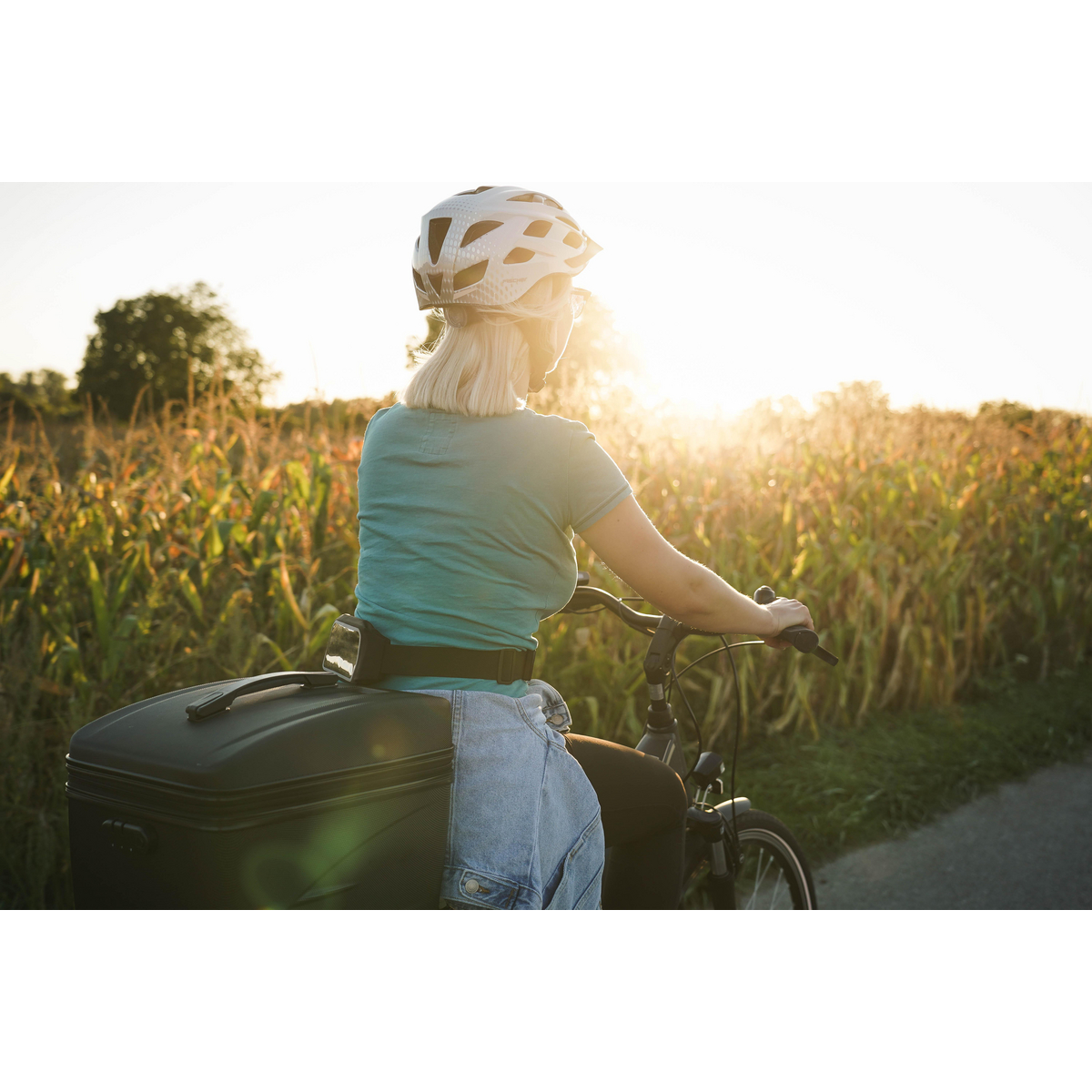 Frau auf dem Fahrrad, blondes Haar, Helm, fährt in der Nähe eines Feldes, Sonne im Hintergrund.