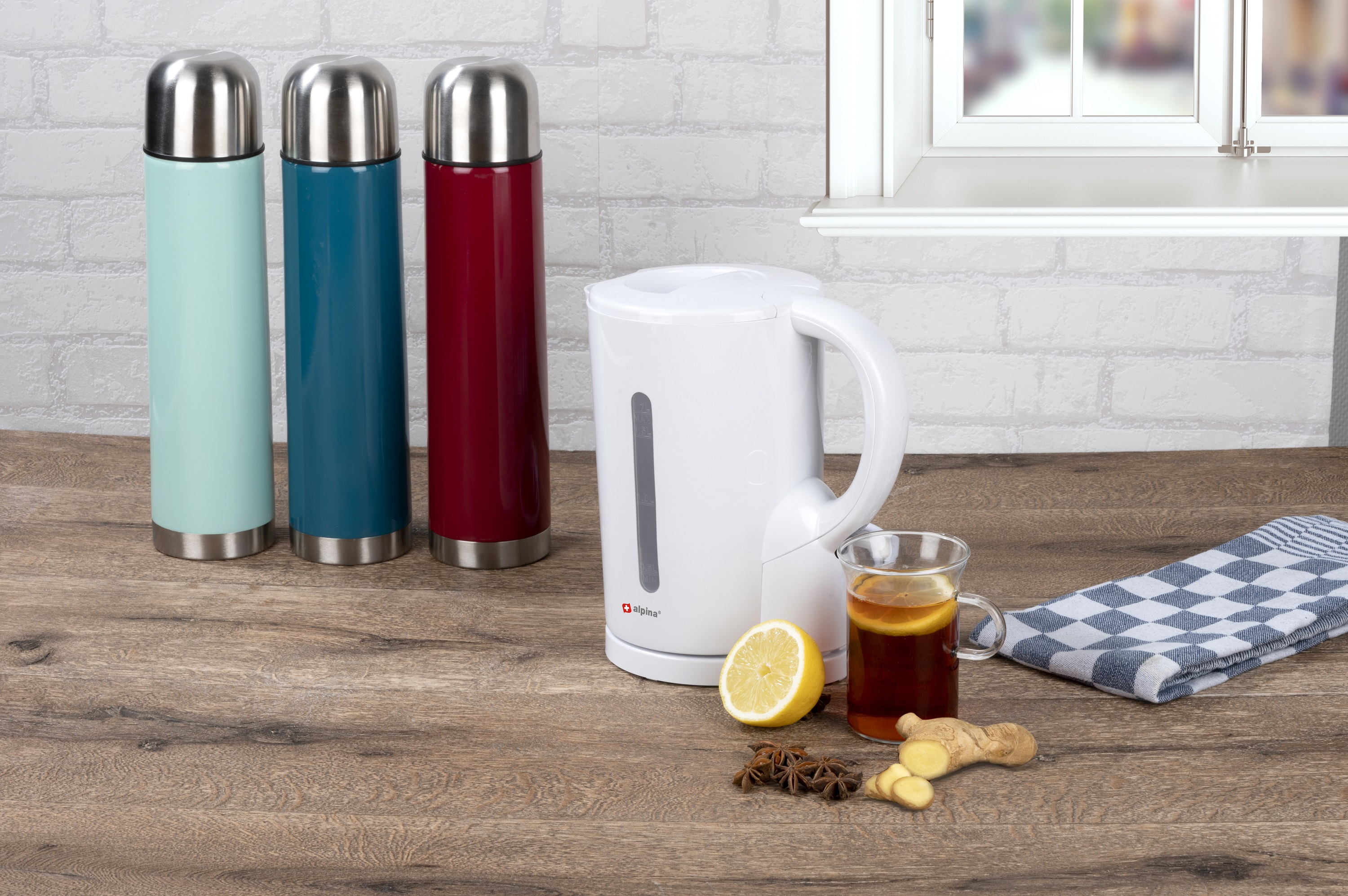 Drei farbige Thermoskannen und ein Wasserkocher mit Tasse Tee und Zutaten auf dem Tisch.