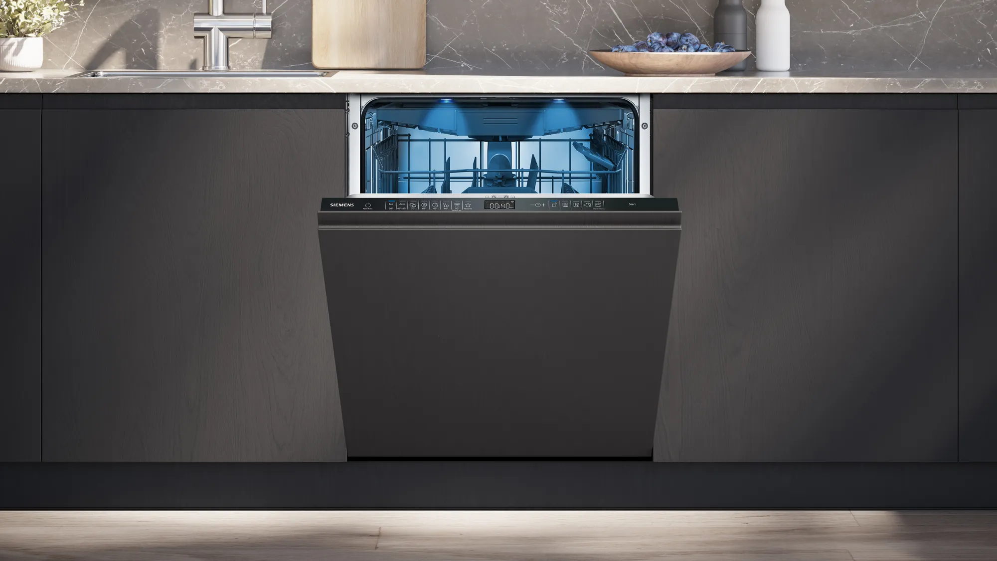 Lavastoviglie con porta aperta. Interno illuminato, mobili in legno scuro sullo sfondo, piano di lavoro con frutta.