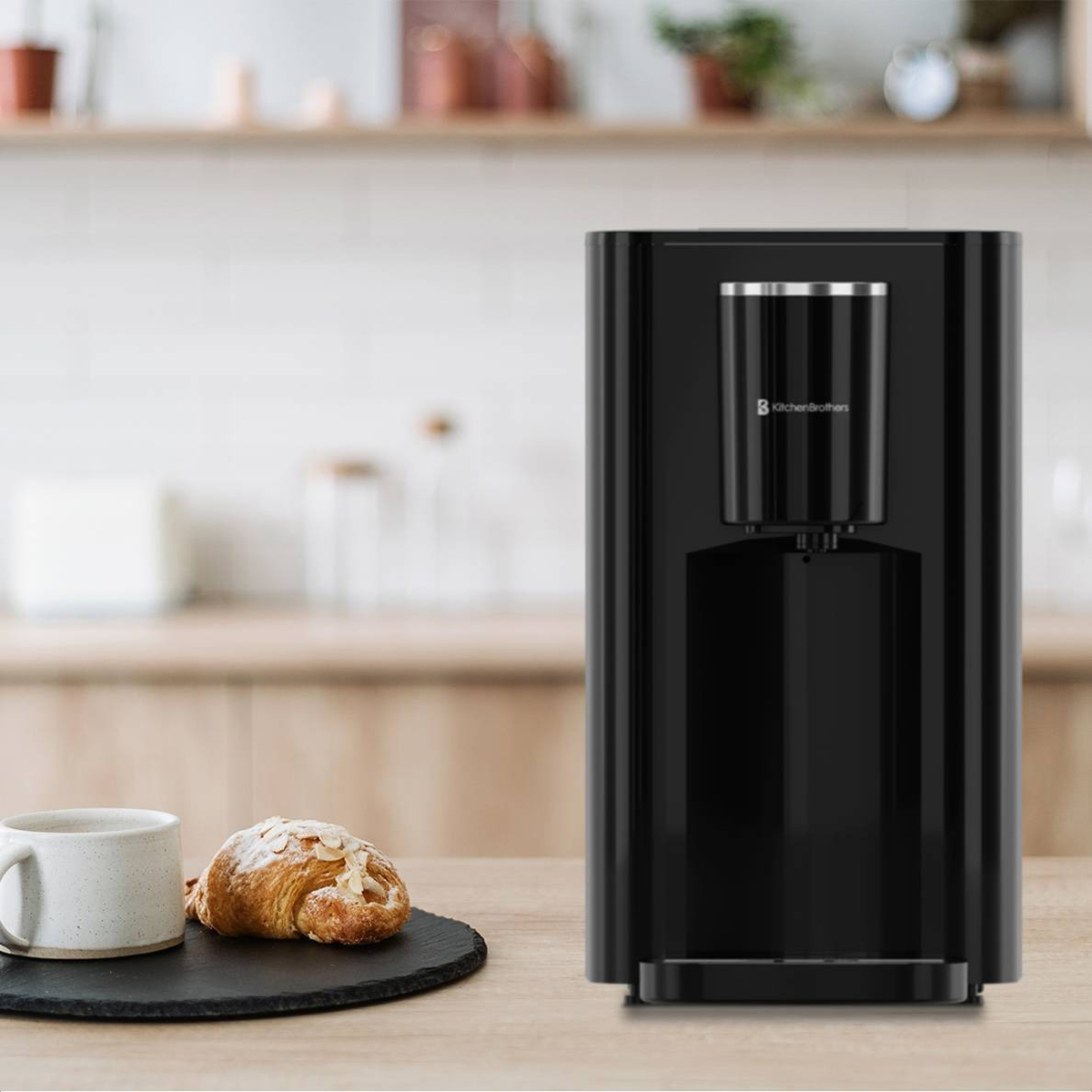 Zwarte waterdispenser met een kopje en croissant op een houten tafel, met een wazige keukenachtergrond.